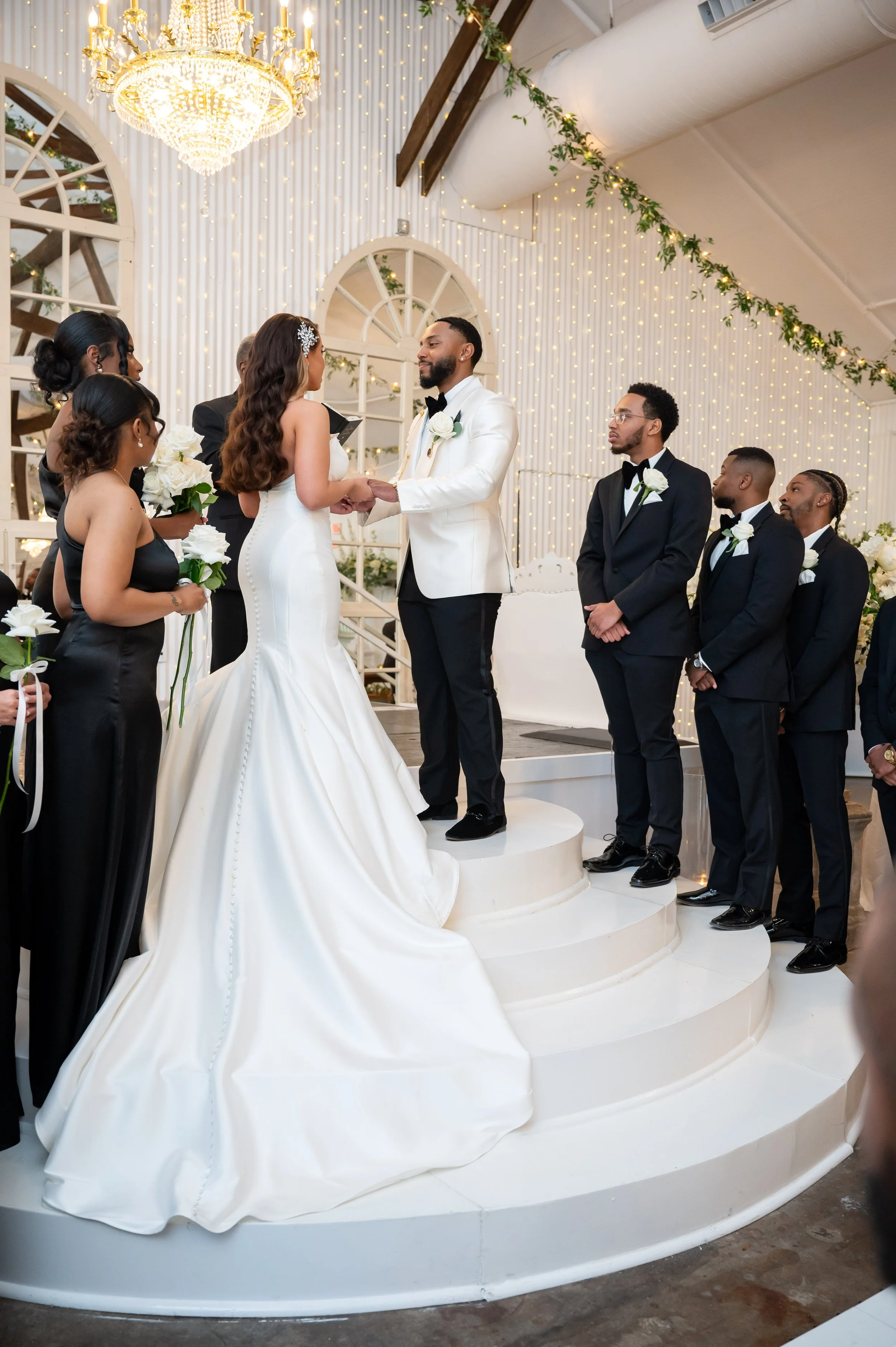 Bride and groom exchange vows on a white circular wedding stage, surrounded by bridesmaids and groomsmen in formal attire in a decorated wedding venue with string lights, a chandelier, and greenery.