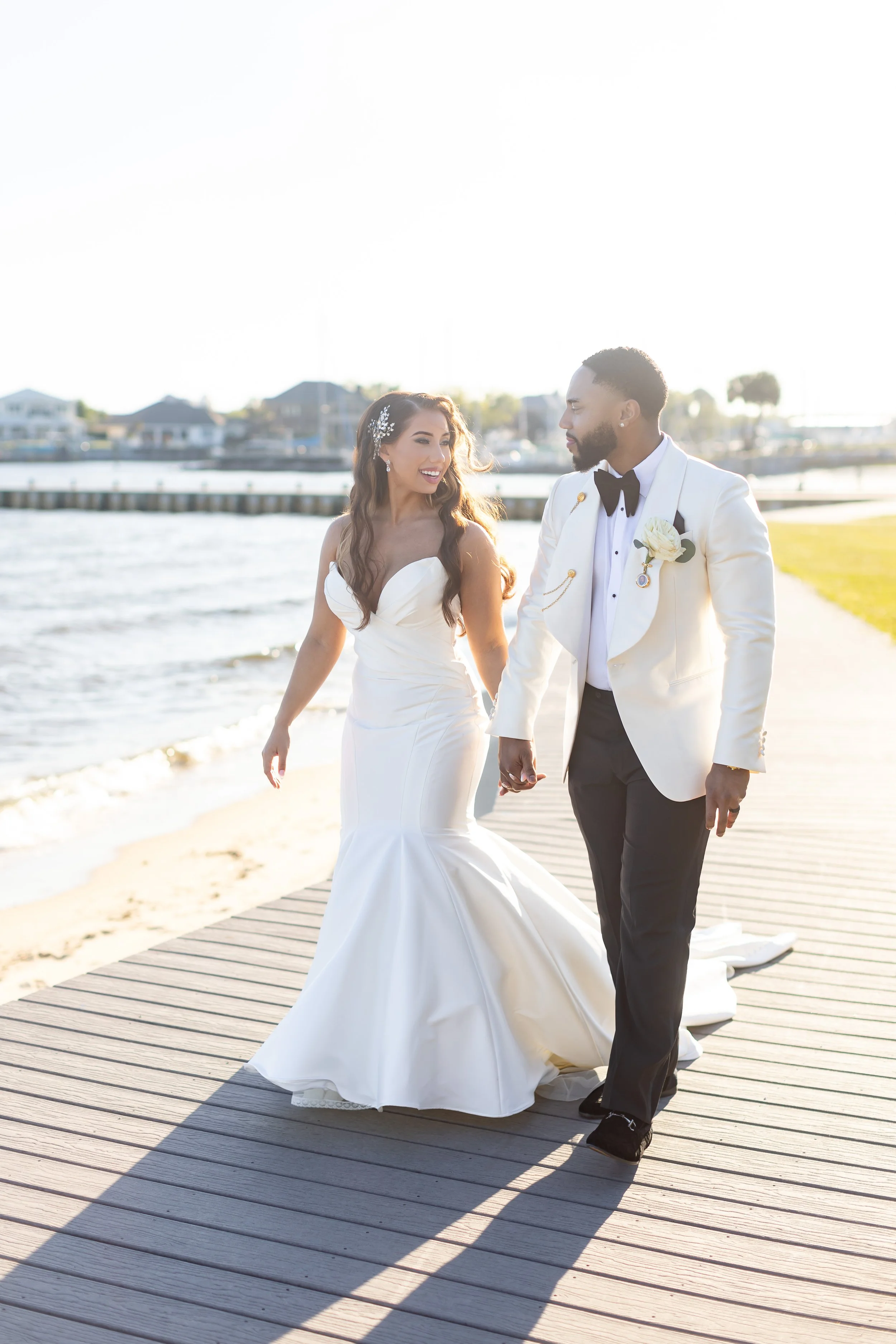 Bride and groom holding hands, walking on a dock beside the water during sunset, dressed in wedding attire in a Pensacola Wedding Venue.