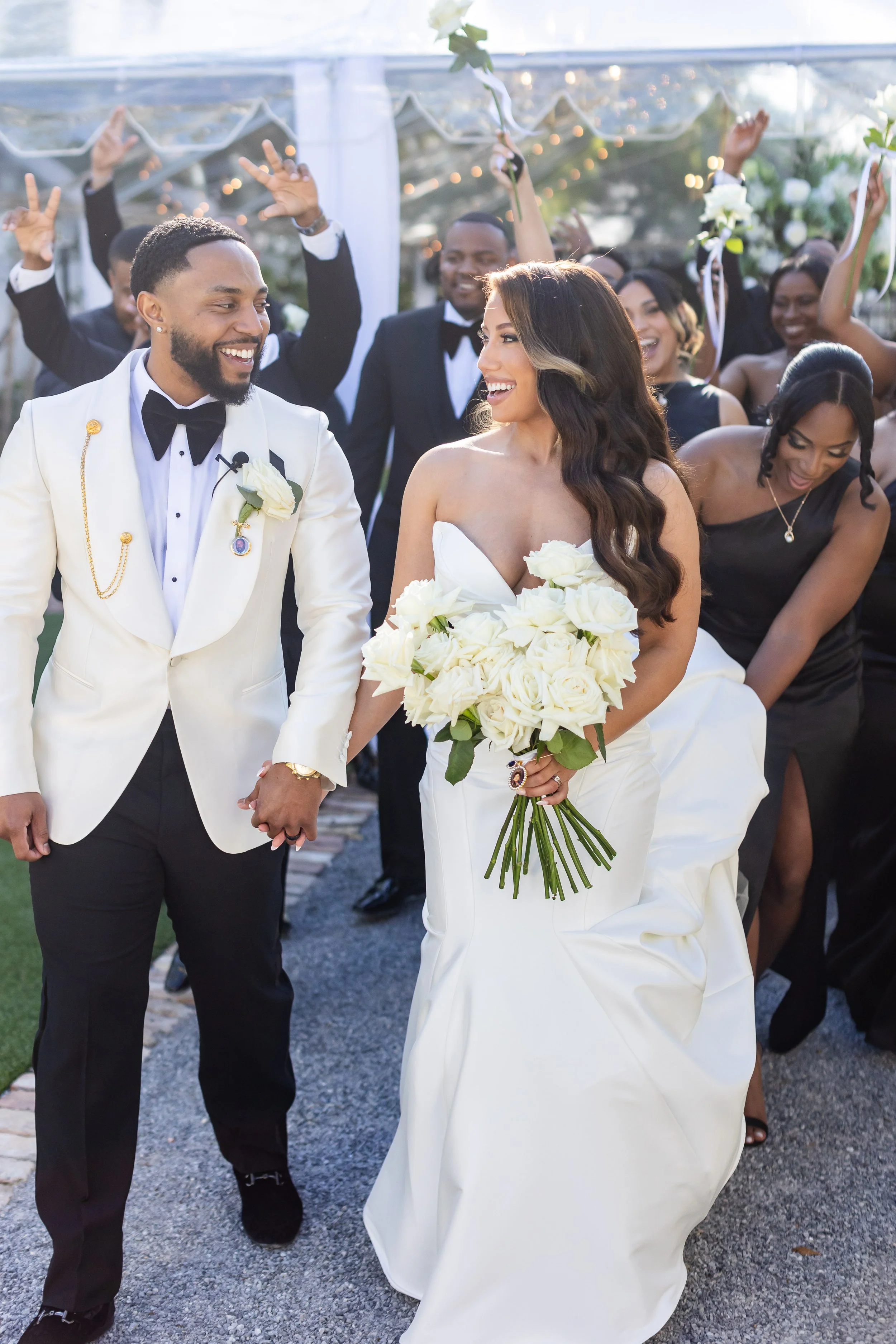 A newlywed couple walking hand-in-hand with friends celebrating behind them at an outdoor wedding ceremony. The bride is holding a bouquet of white roses and wearing a strapless white wedding gown. The groom is dressed in a white tuxedo jacket with b