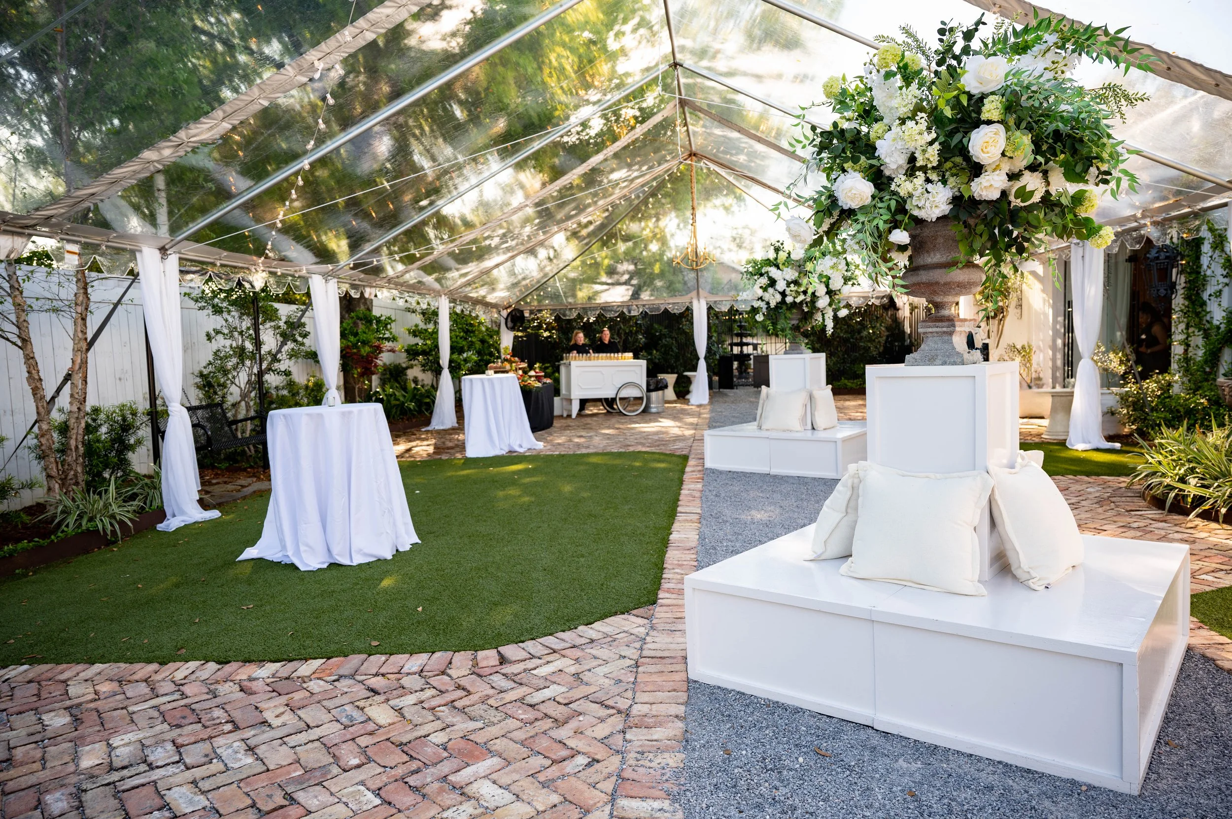 A decorated outdoor event space with a transparent tent, white fabric drapes, elegant white furniture, floral arrangements, and string lights, set on a brick and grass surface in a Pensacola Wedding Venue.