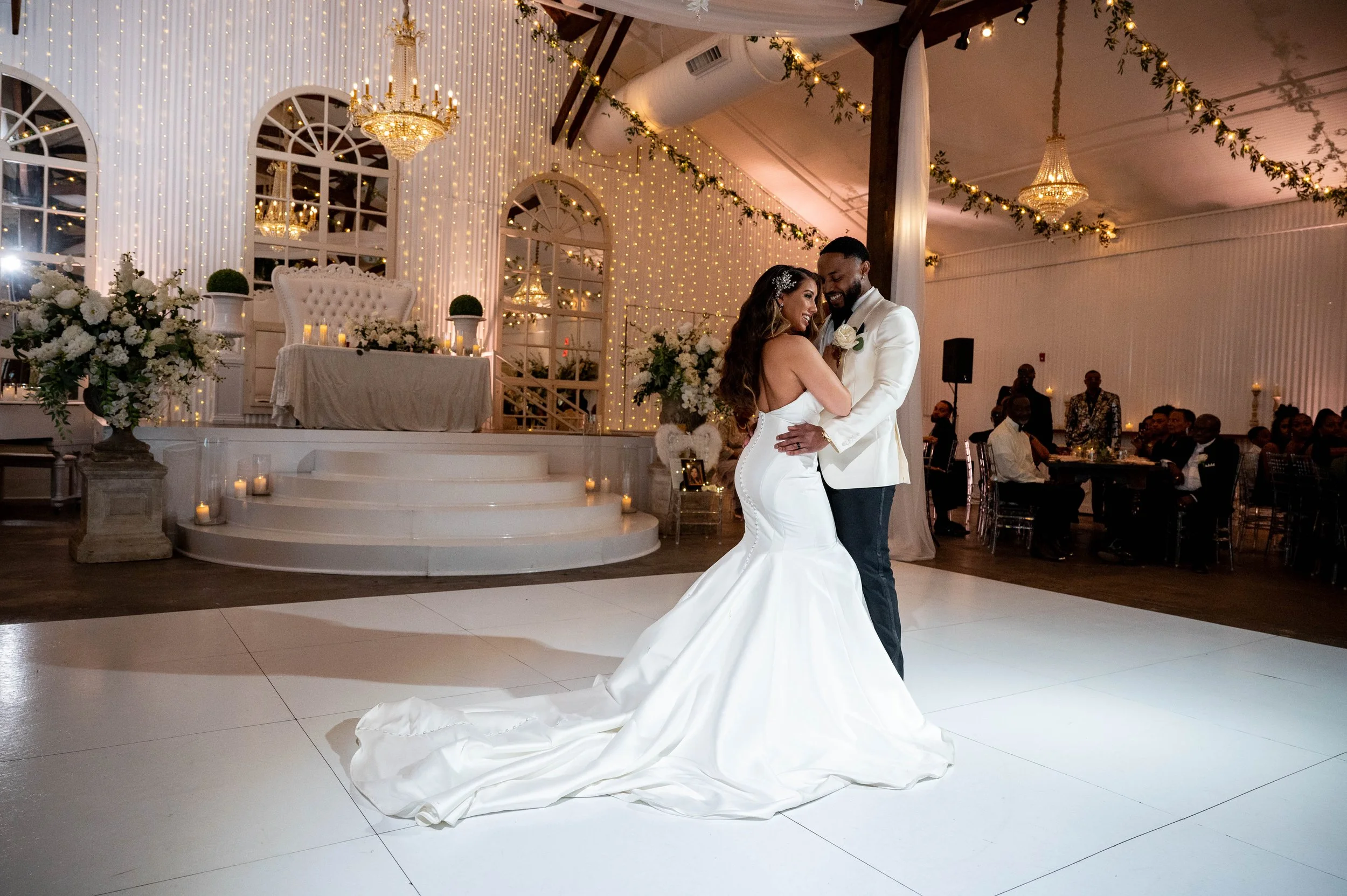 A bride and groom share a dance at their wedding reception. The bride wears a white strapless wedding gown with a long train, and the groom wears a white tuxedo jacket with black pants. Elegant decorations with flowers, candles, and string lights are
