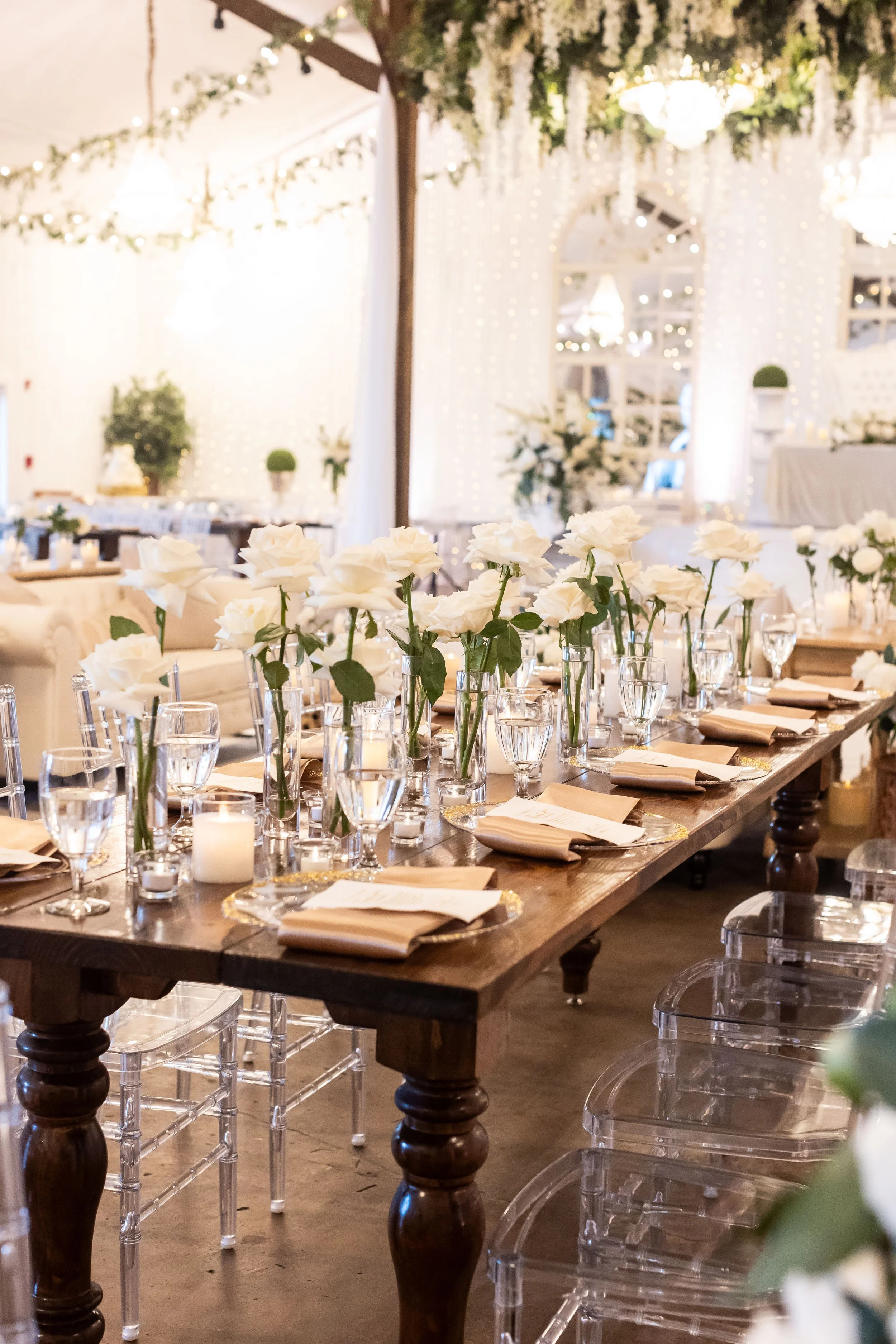 Elegant event table setup with white roses in vases, candles, glasses, and neatly folded napkins in a decorated venue with string lights and white drapery.