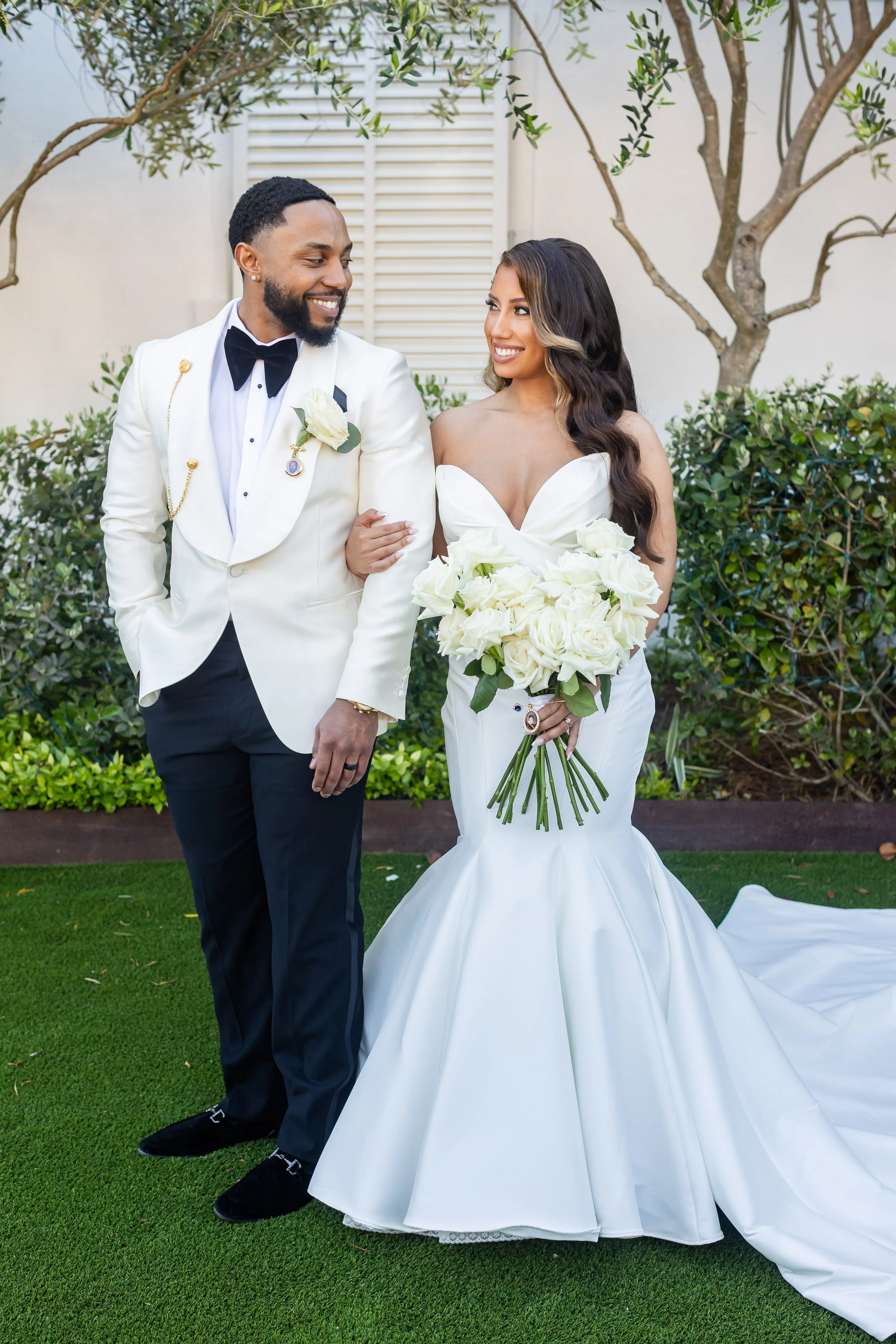 A bride and groom stand outdoors, smiling and looking at each other. The bride wears a white strapless wedding gown and holds a bouquet of white roses. The groom wears a white tuxedo jacket with black pants, a black bow tie, and a white boutonnière. 