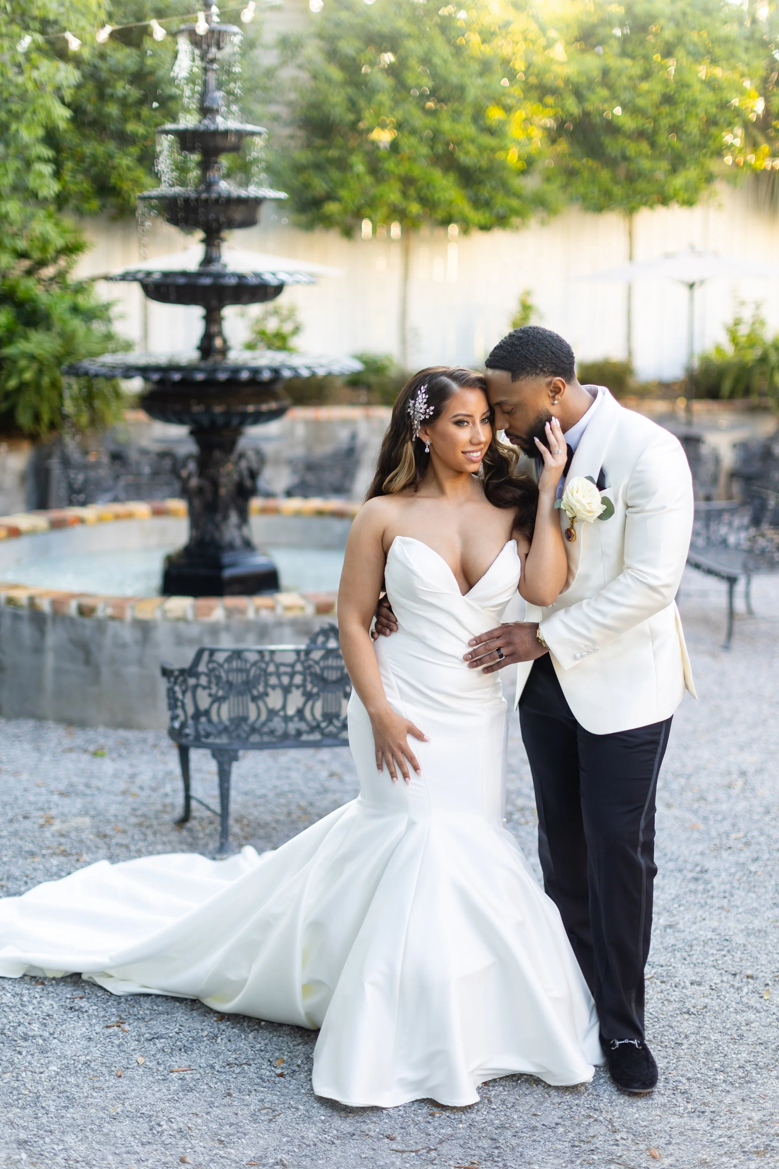 Bride and groom embrace outdoors at a wedding venue with a fountain and green trees, dressed in wedding attire.