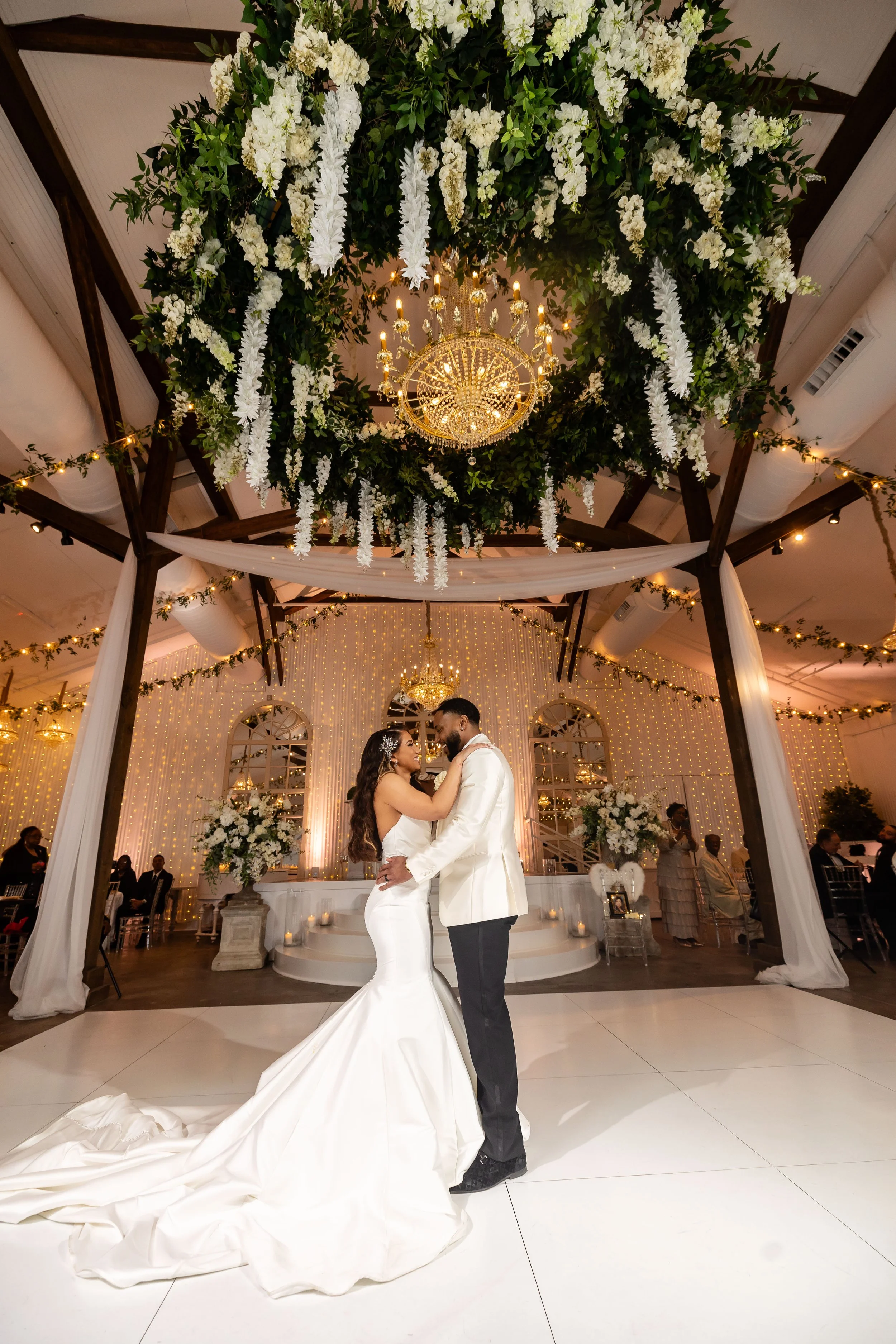 A bride and groom share a dance under an elegant hanging floral chandelier at their wedding reception, decorated with white flowers, greenery, fairy lights, and draped fabric.