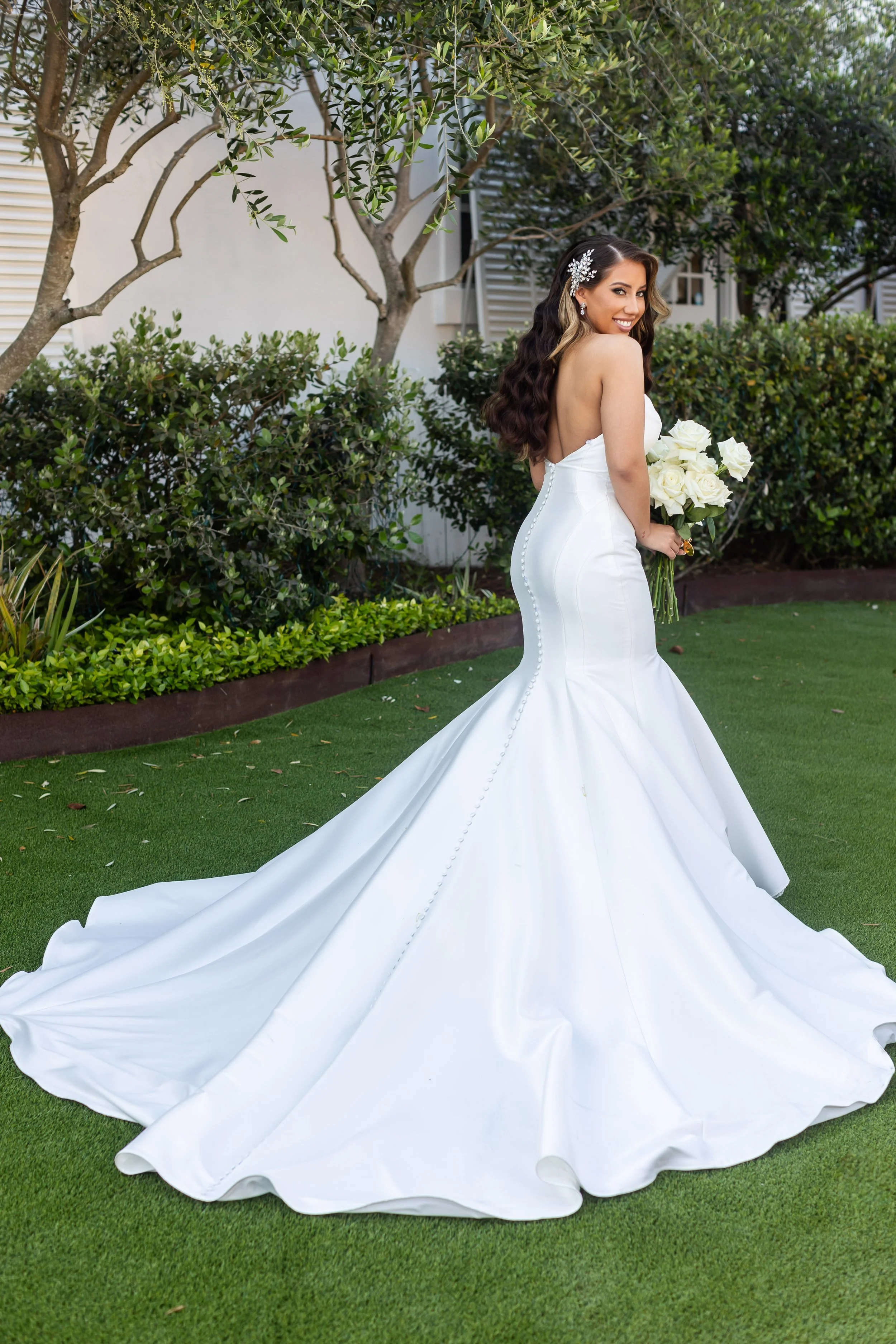 A bride in a strapless white wedding gown with a long train standing outdoors on a grassy area, holding a bouquet of white roses, and smiling