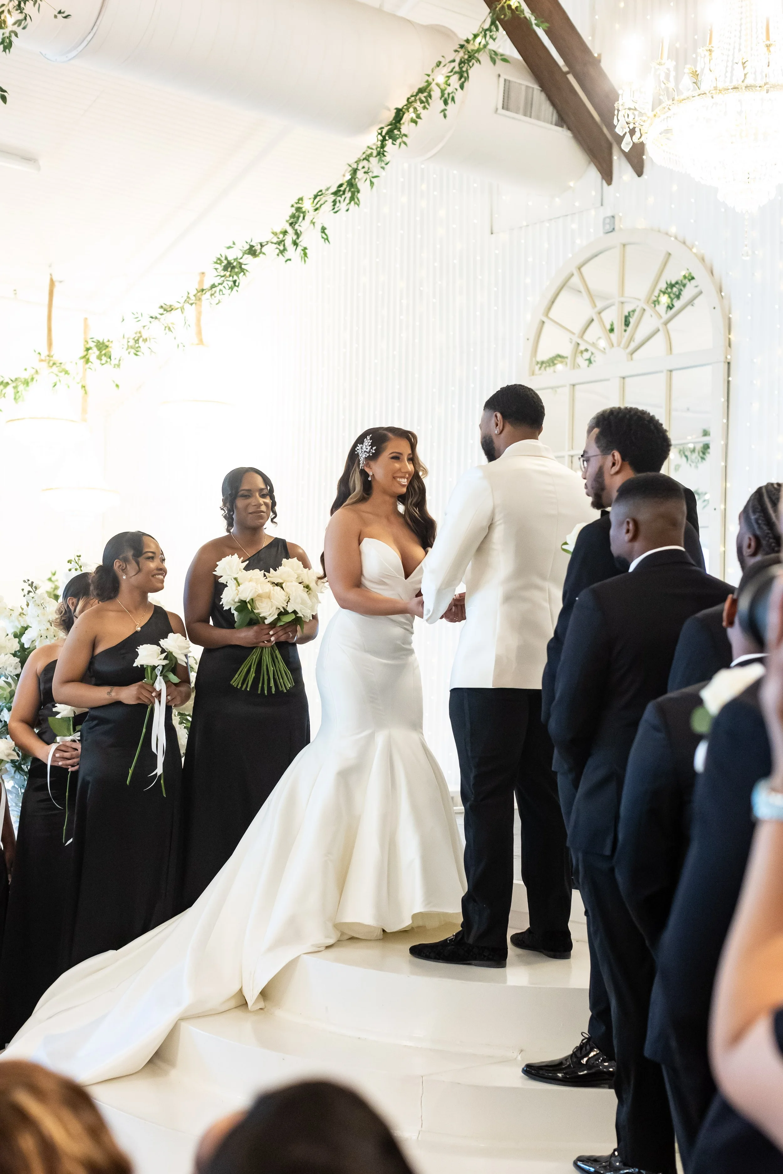 The image shows a wedding ceremony with a bride and groom standing at the altar, holding hands and exchanging vows. The bride is wearing a strapless white wedding gown, and the groom is dressed in a white jacket and dark pants. Bridesmaids and grooms