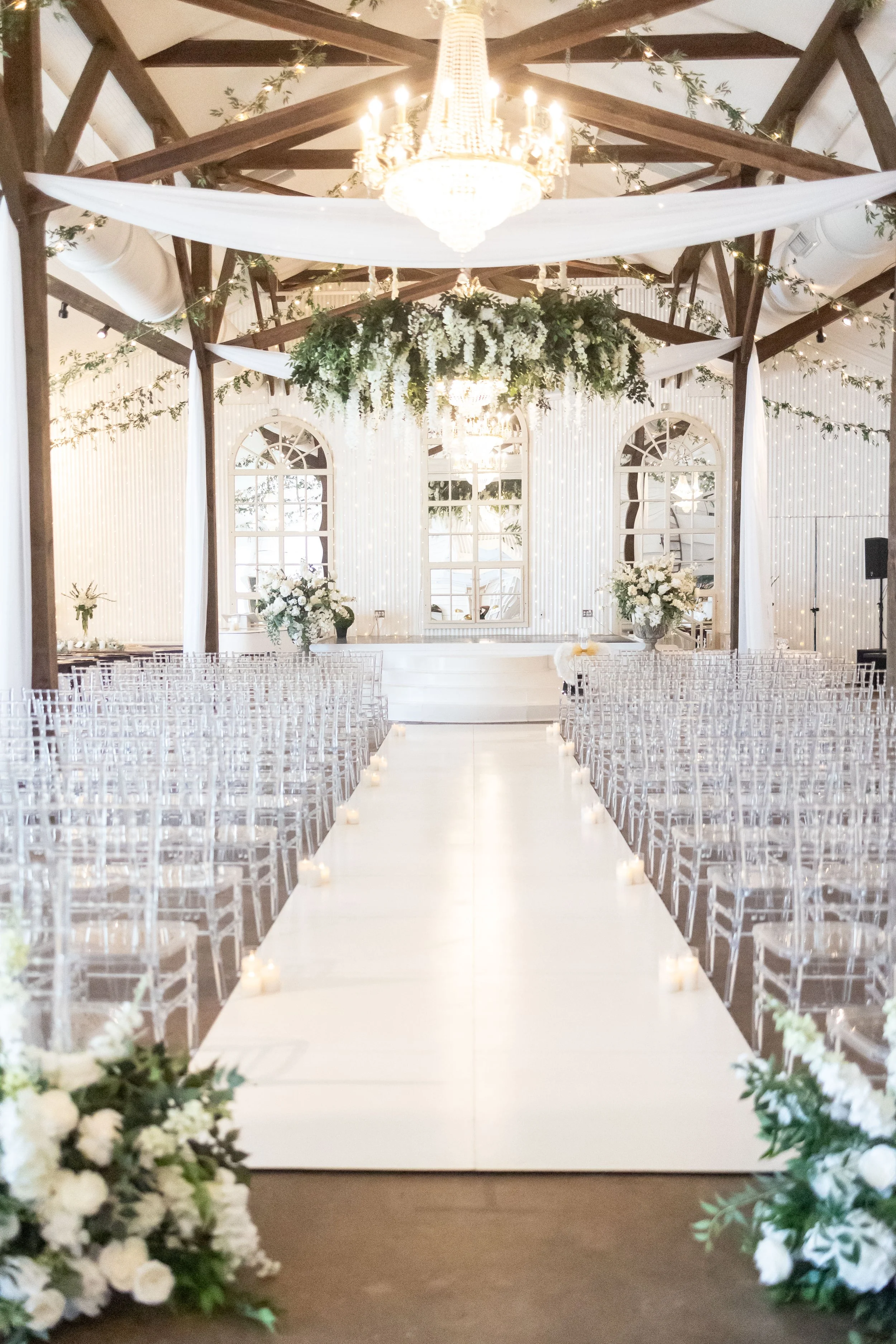 Elegant wedding ceremony setup inside a decorated venue with a white aisle, floral arrangements, and chandeliers hanging from a wooden ceiling.