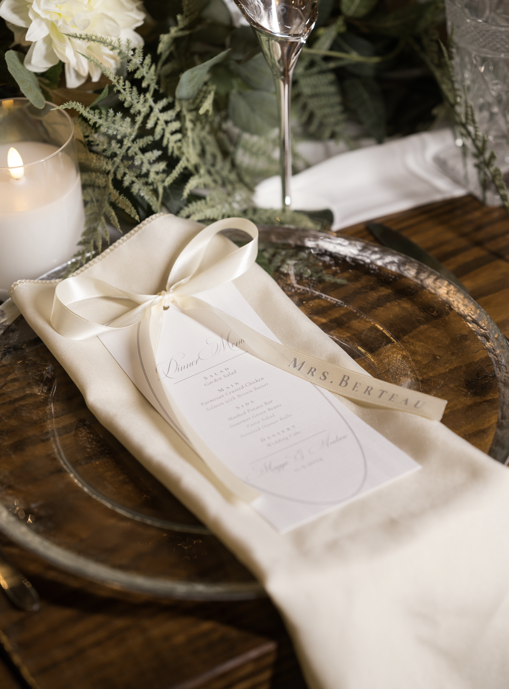 Elegant table setting with a wedding menu, cream-colored napkin with a ribbon, surrounded by greenery, a lit candle, and a wine glass.