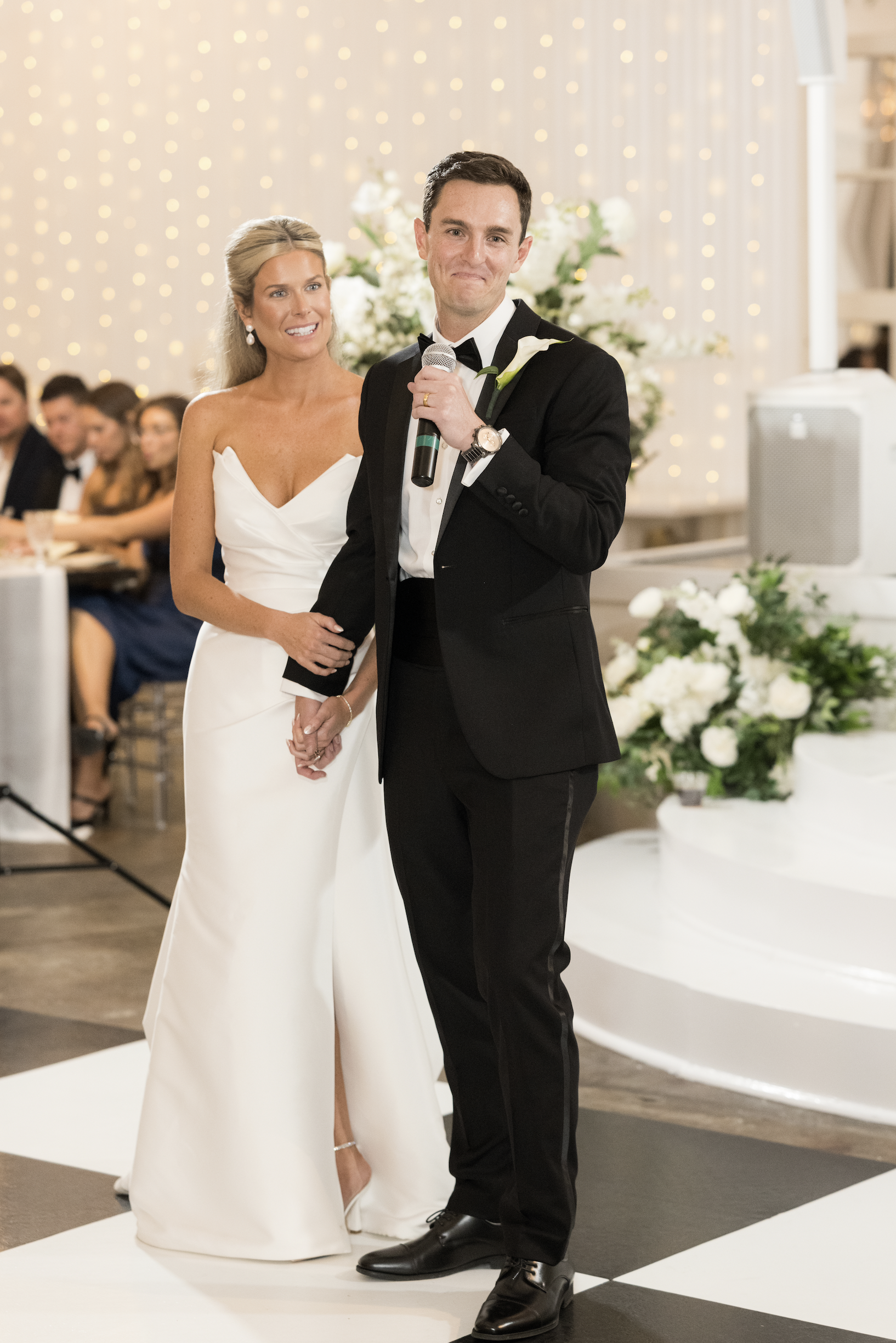 A bride and groom standing together during their wedding reception, with the groom holding a microphone, in front of guests and floral decorations.