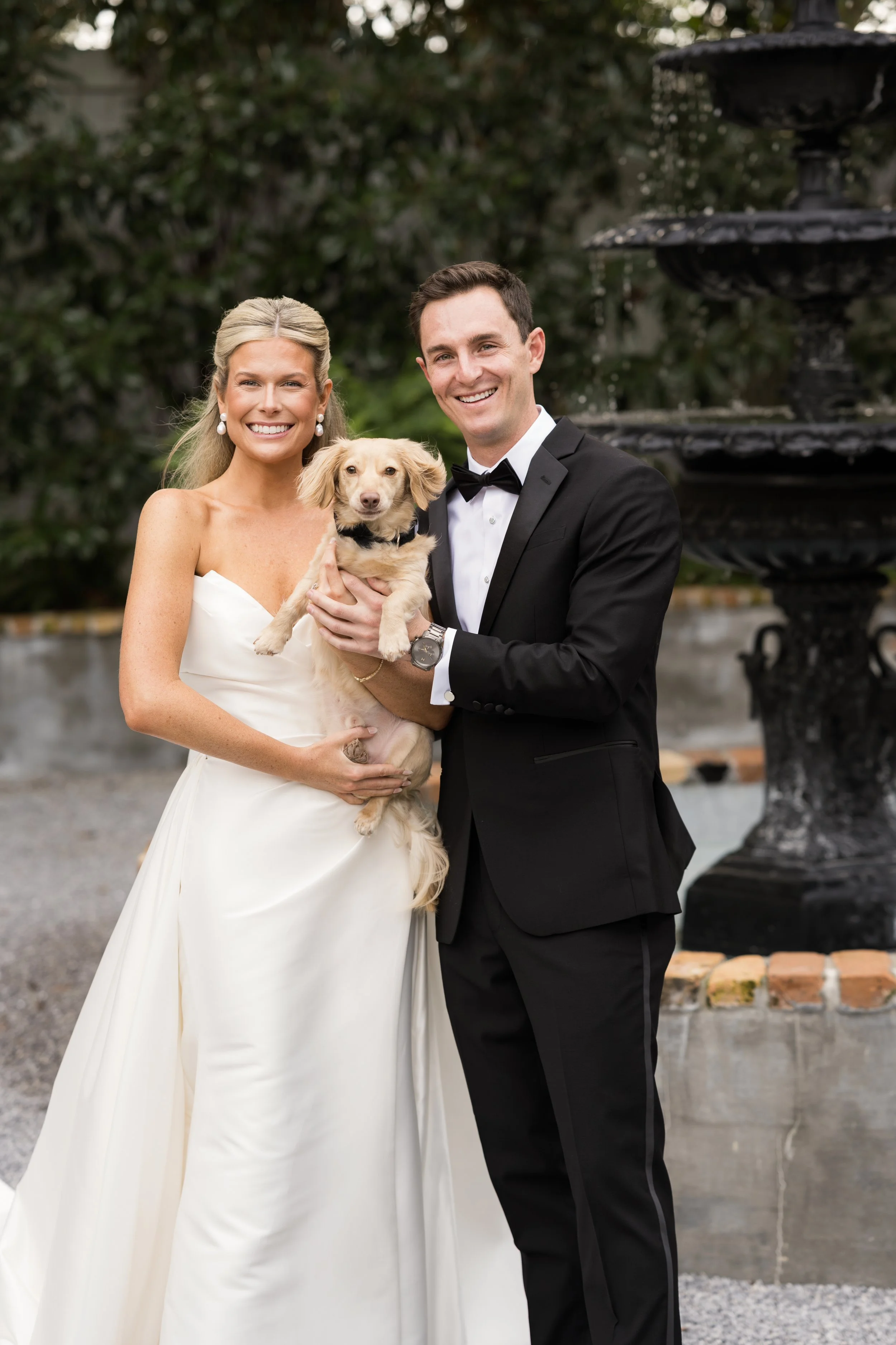A bride and groom dressed in wedding attire, holding a small dog, smiling outdoors with a fountain in the background of a Pensacola Wedding Venue.