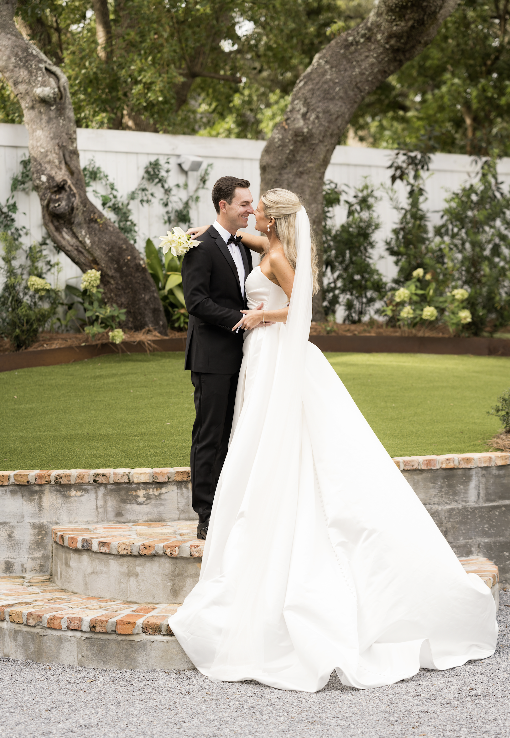 A bride and groom stand close together on a small brick staircase outdoors, smiling at each other, with green trees and shrubs in the background.