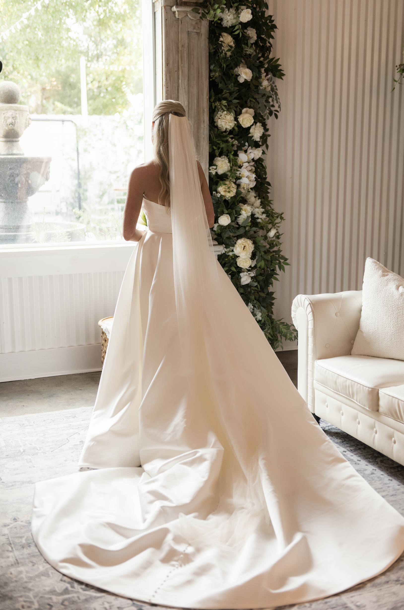 Bride in a strapless satin wedding gown, standing by a window with sunlight, with a long veil, next to a floral wall, in a rustic indoor setting.