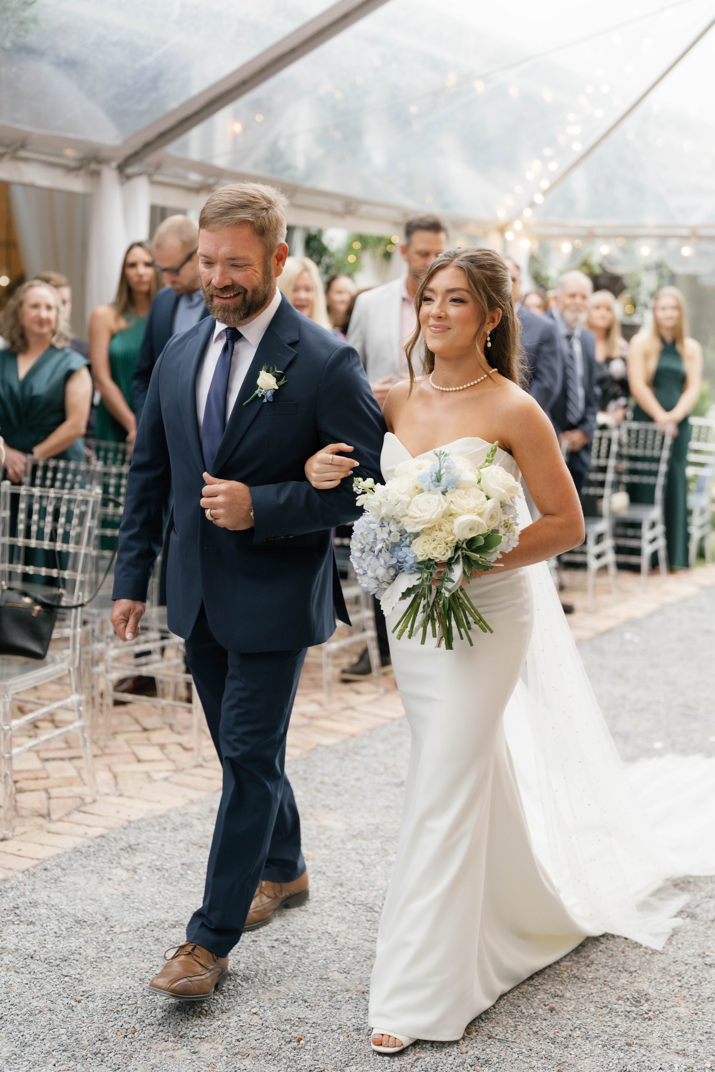 A bride in a white wedding gown carrying a flower bouquet walks arm-in-arm with a man in a navy suit, as guests look on at a wedding reception under a transparent tent.