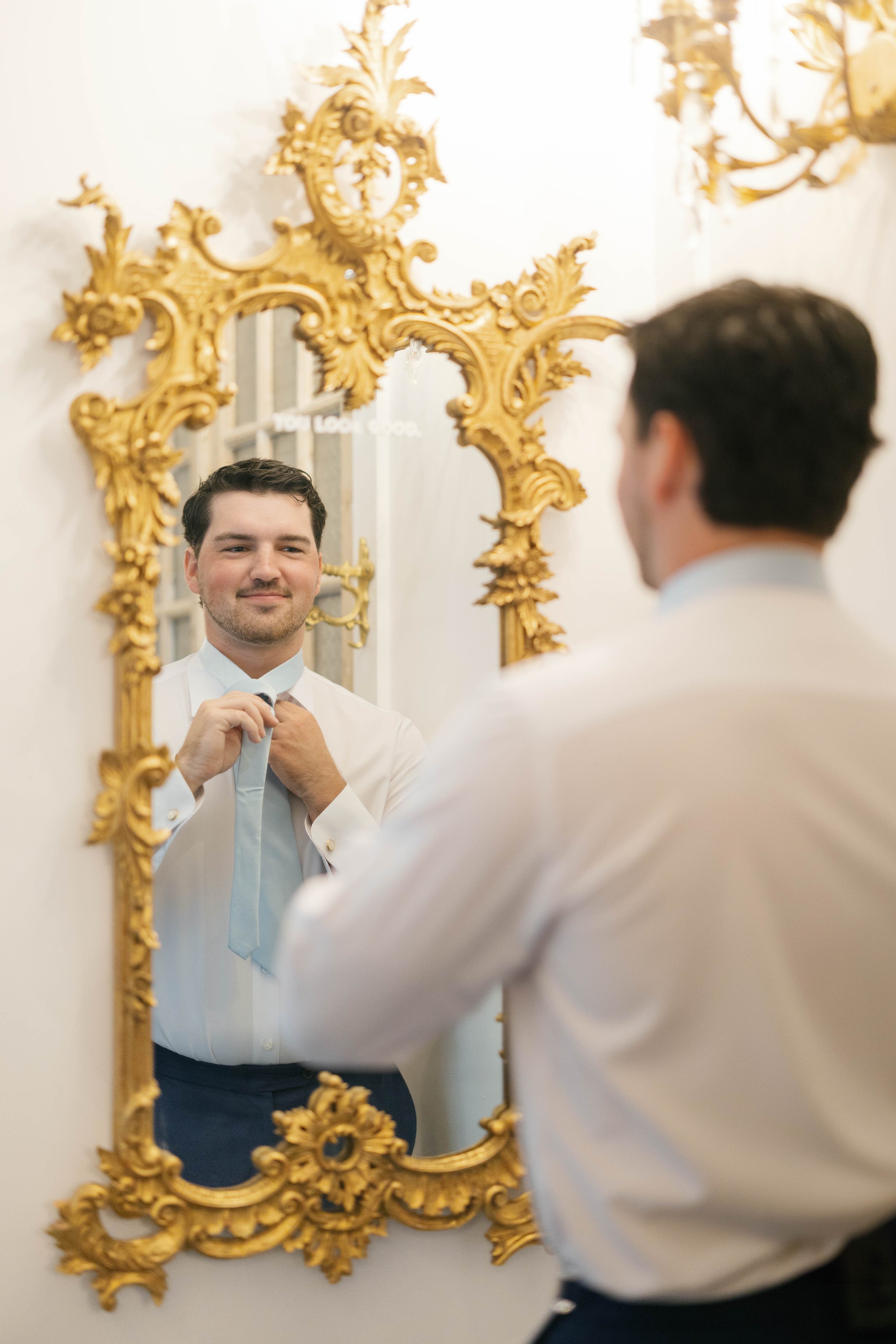 A man is tying a light blue tie while looking at himself in an ornate gold-framed mirror.