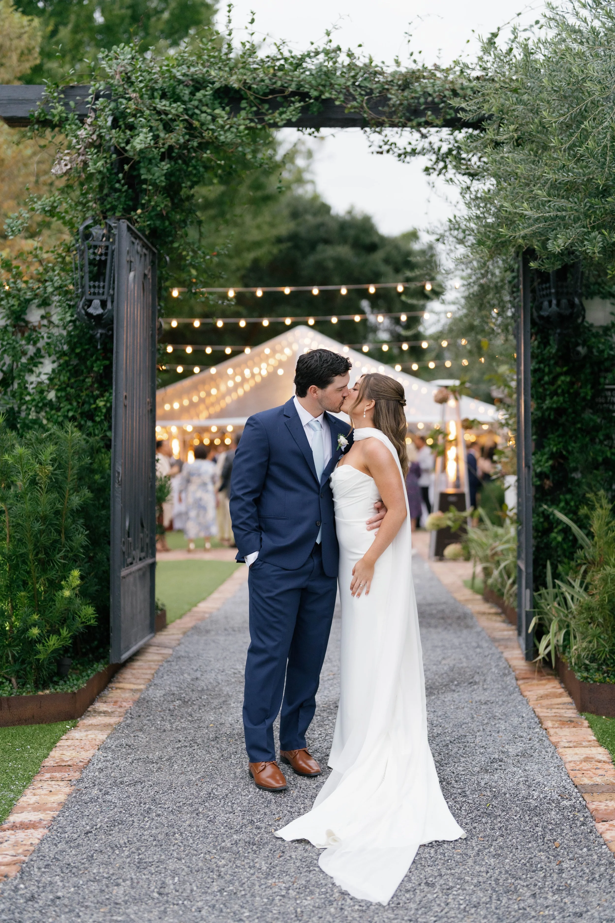 A newlywed couple sharing a kiss at their outdoor wedding reception under string lights, with guests and a white tent in the background of a Pensacola Wedding Venue.