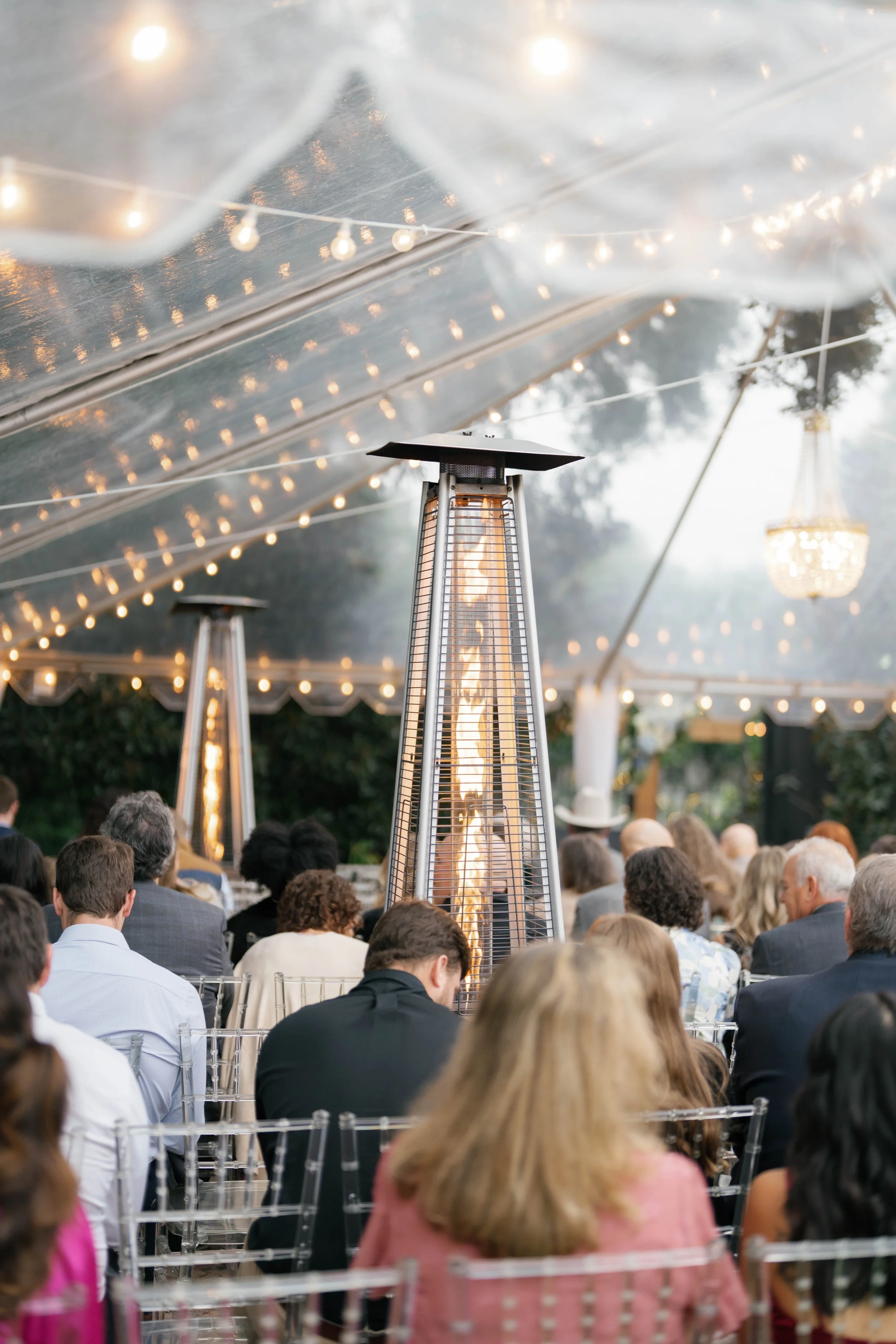 Outdoor event under a clear tent with string lights, featuring patio heaters, with a seated crowd watching a performance or speaker at a Pensacola Wedding Venue.