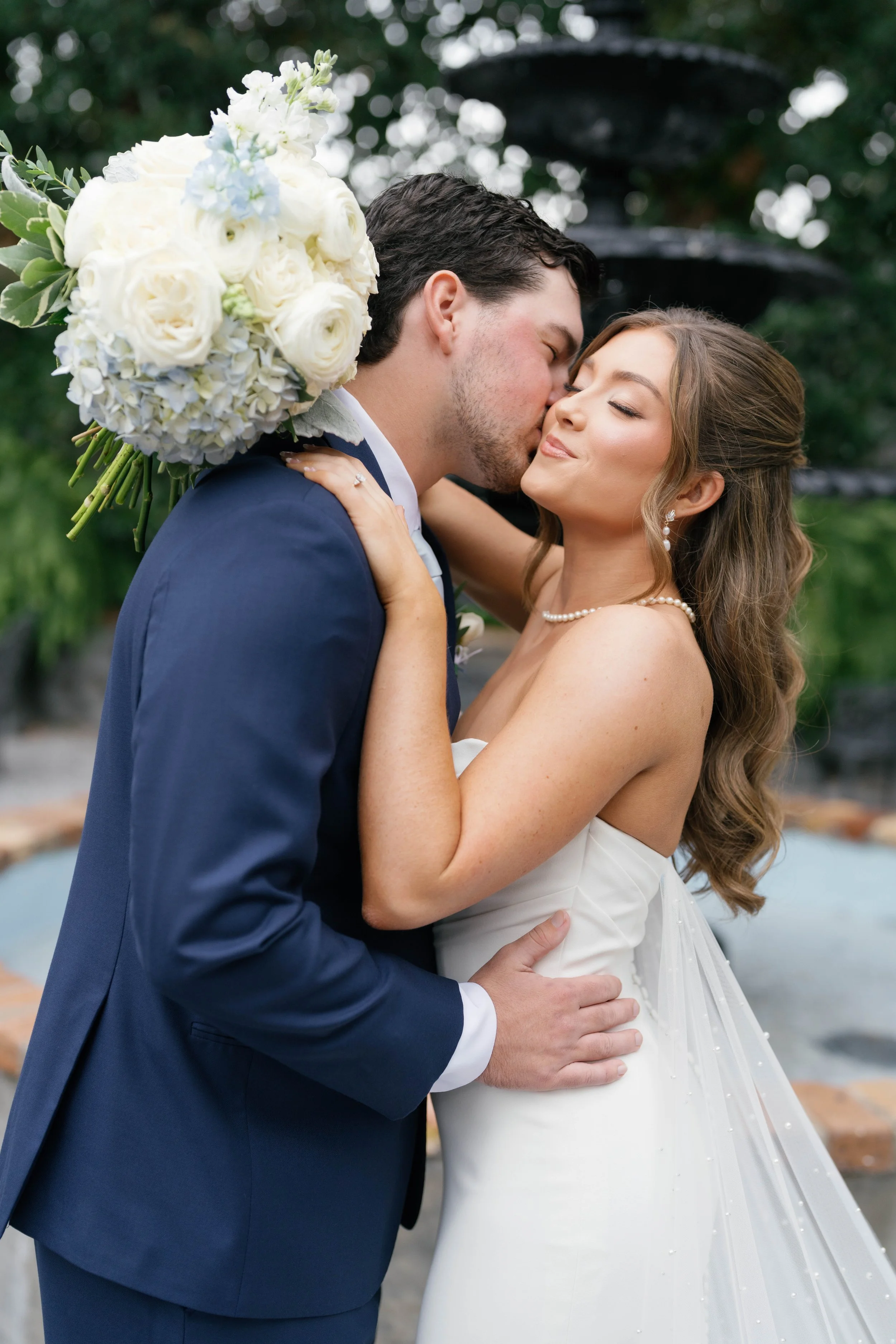 A newlywed couple sharing a kiss outdoors, with the groom holding a large bouquet of white roses and hydrangeas, while the bride has long wavy hair and wears a strapless white wedding dress, pearl necklace, and earrings in a Pensacola Wedding Venue.