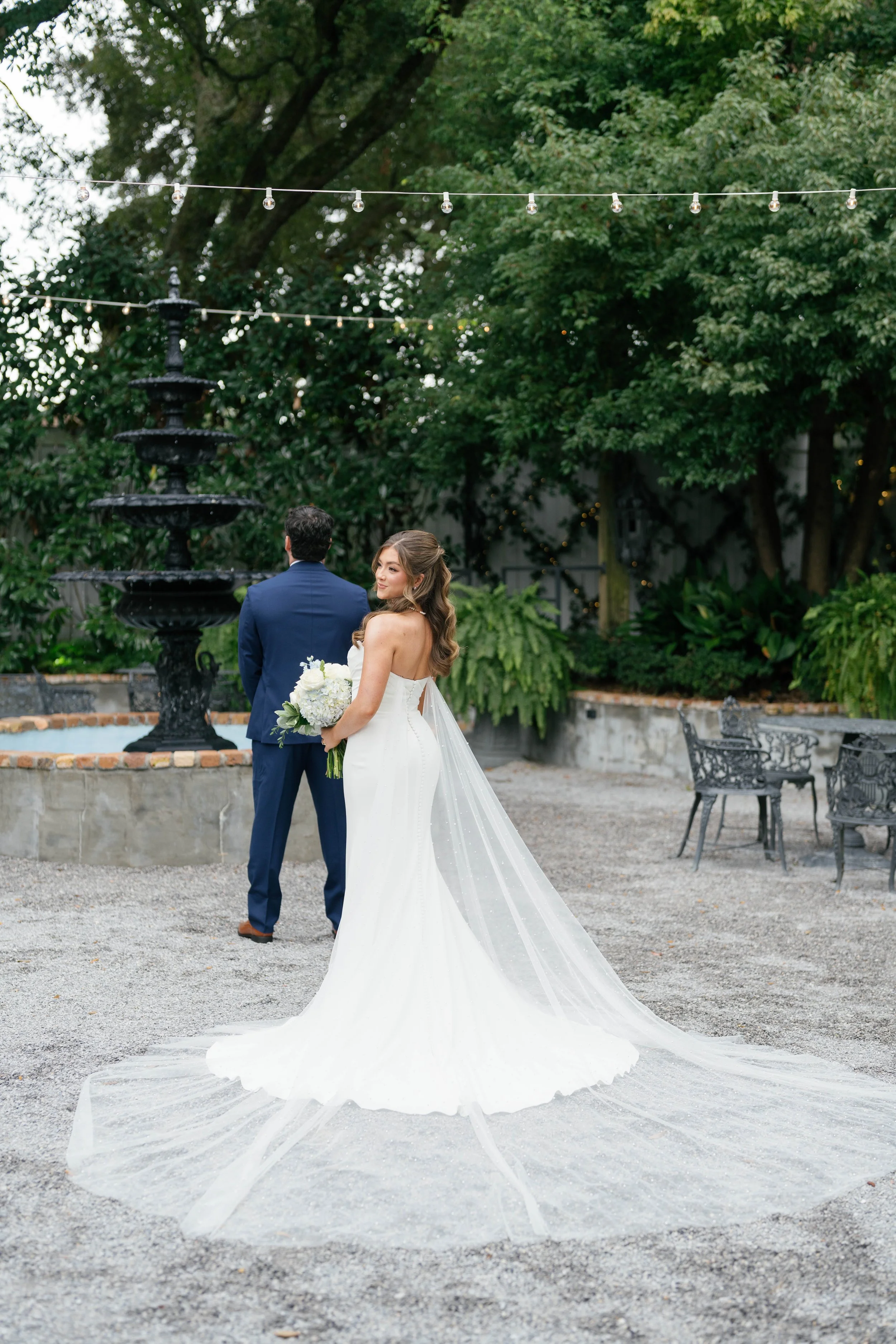 Bride holding a bouquet in a white wedding dress standing outdoors with groom in a blue suit in front of a fountain, surrounded by trees and string lights.