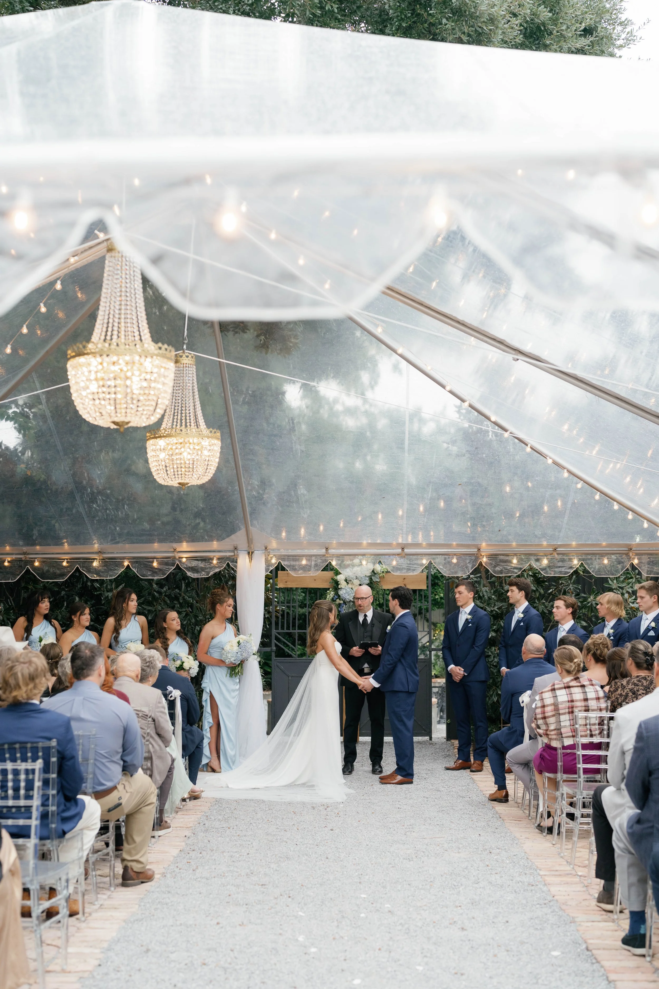 A wedding ceremony taking place outdoors under a transparent tent with chandeliers and string lights, with the bride and groom holding hands, officiant reading, and guests seated around.