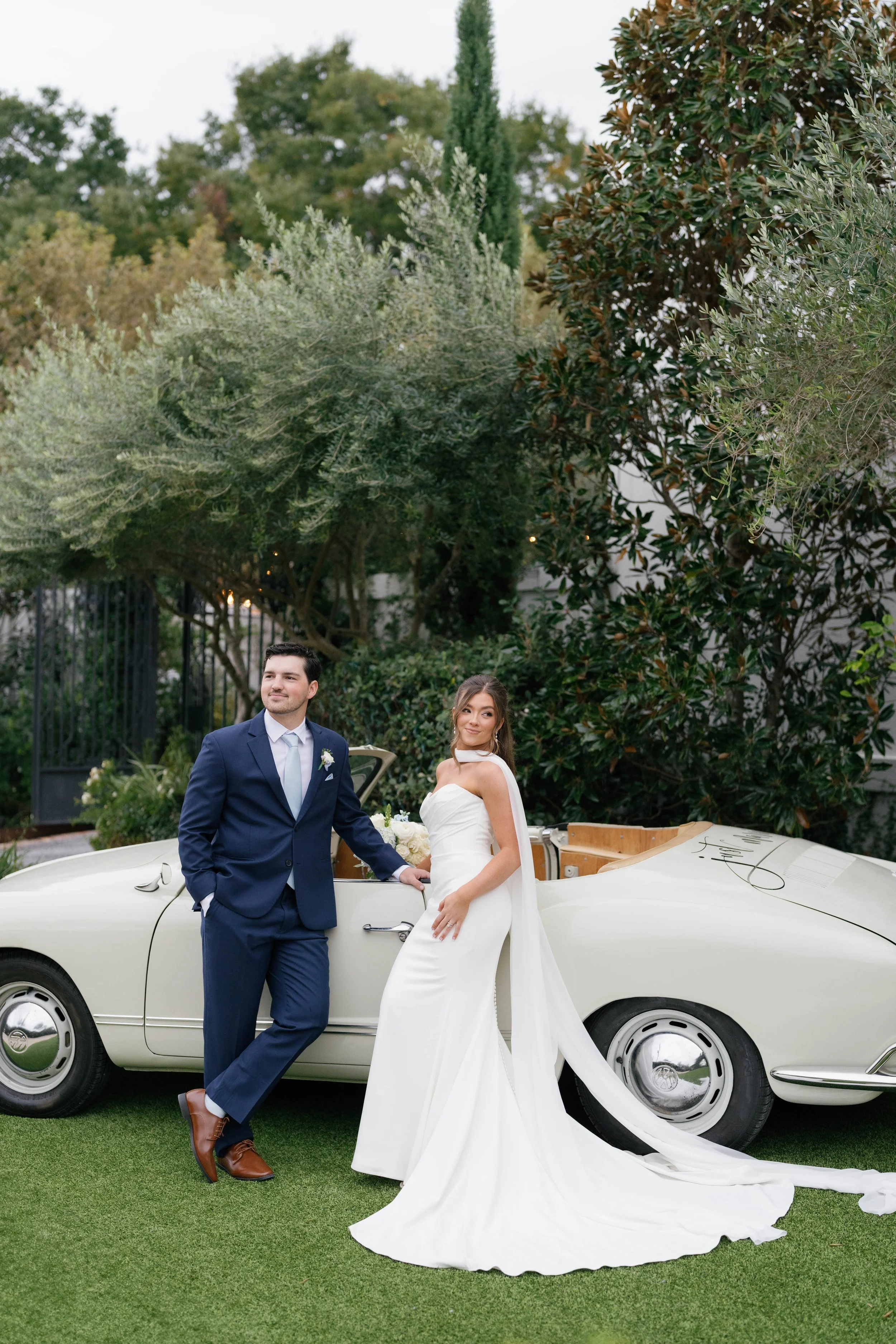 A bride and groom standing next to a vintage white convertible car outdoors, surrounded by trees. The groom is wearing a navy blue suit and light blue tie, while the bride is in a white strapless wedding gown with a long train.