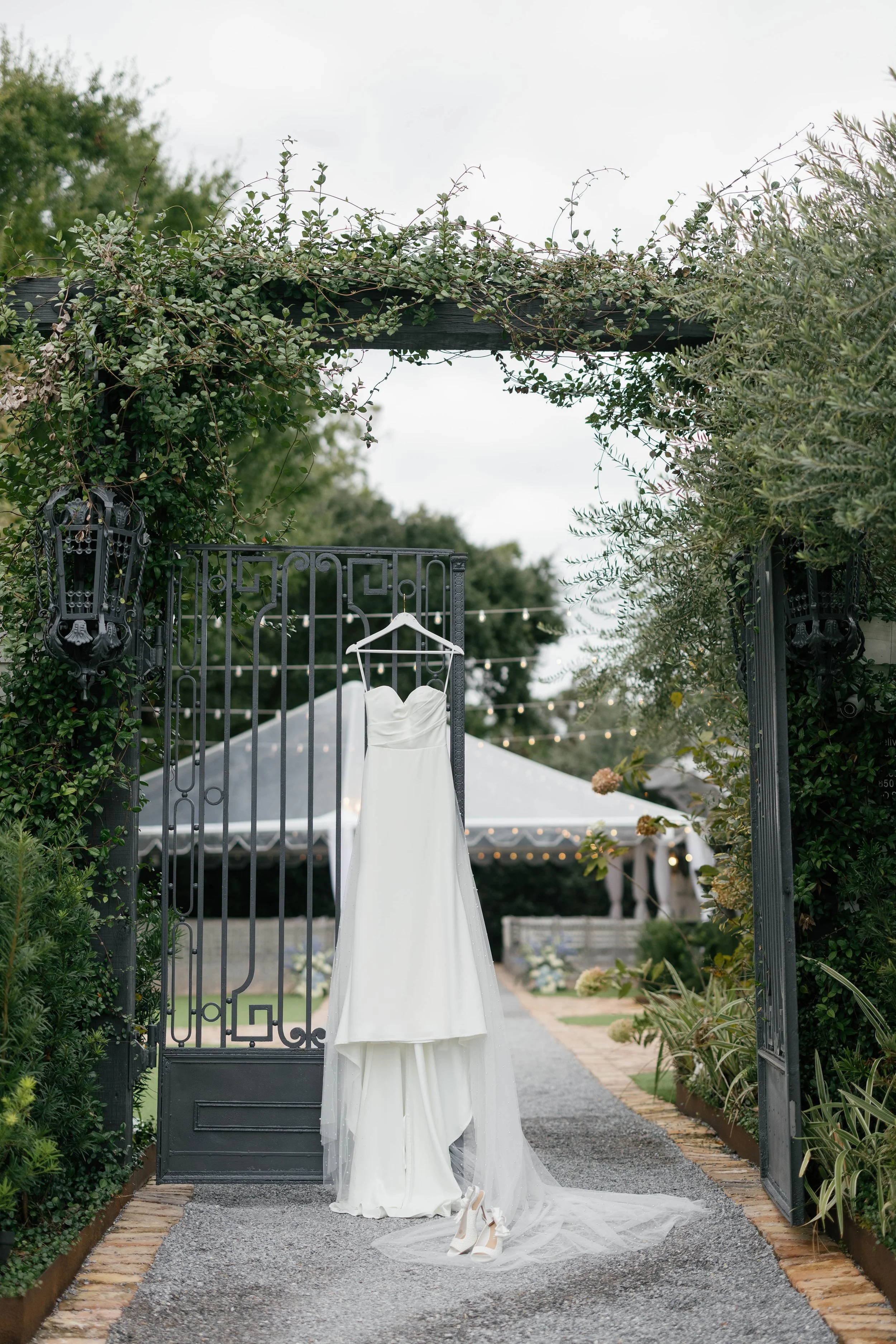 A white wedding dress hanging on a black metal gate under a garden arch with greenery. Wedding shoes placed on the ground in front of the dress, with a tent and string lights in the background.