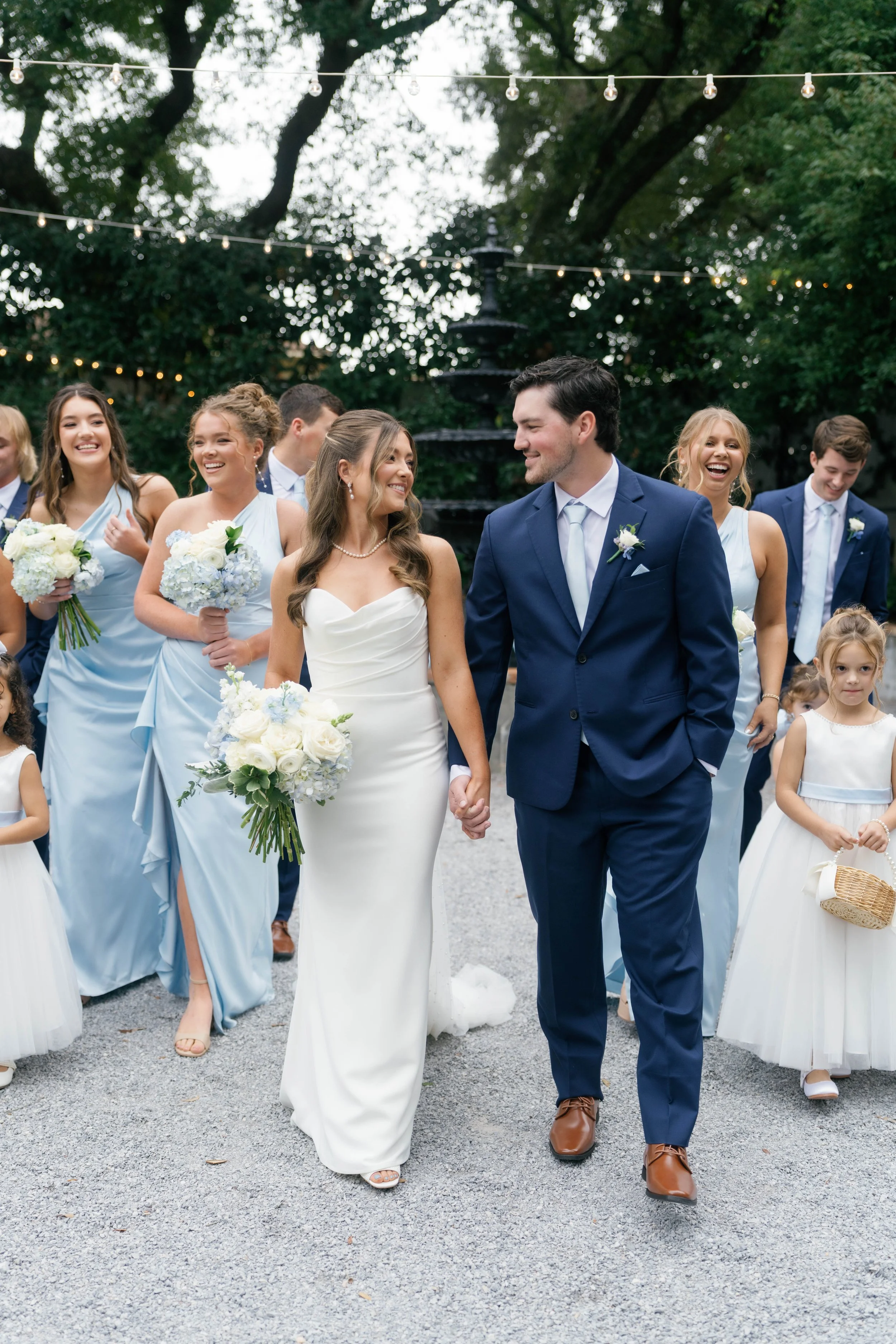 A bride and groom holding hands and smiling, walking with their wedding party including bridesmaids and flower girls, outdoors with string lights and trees in the background.