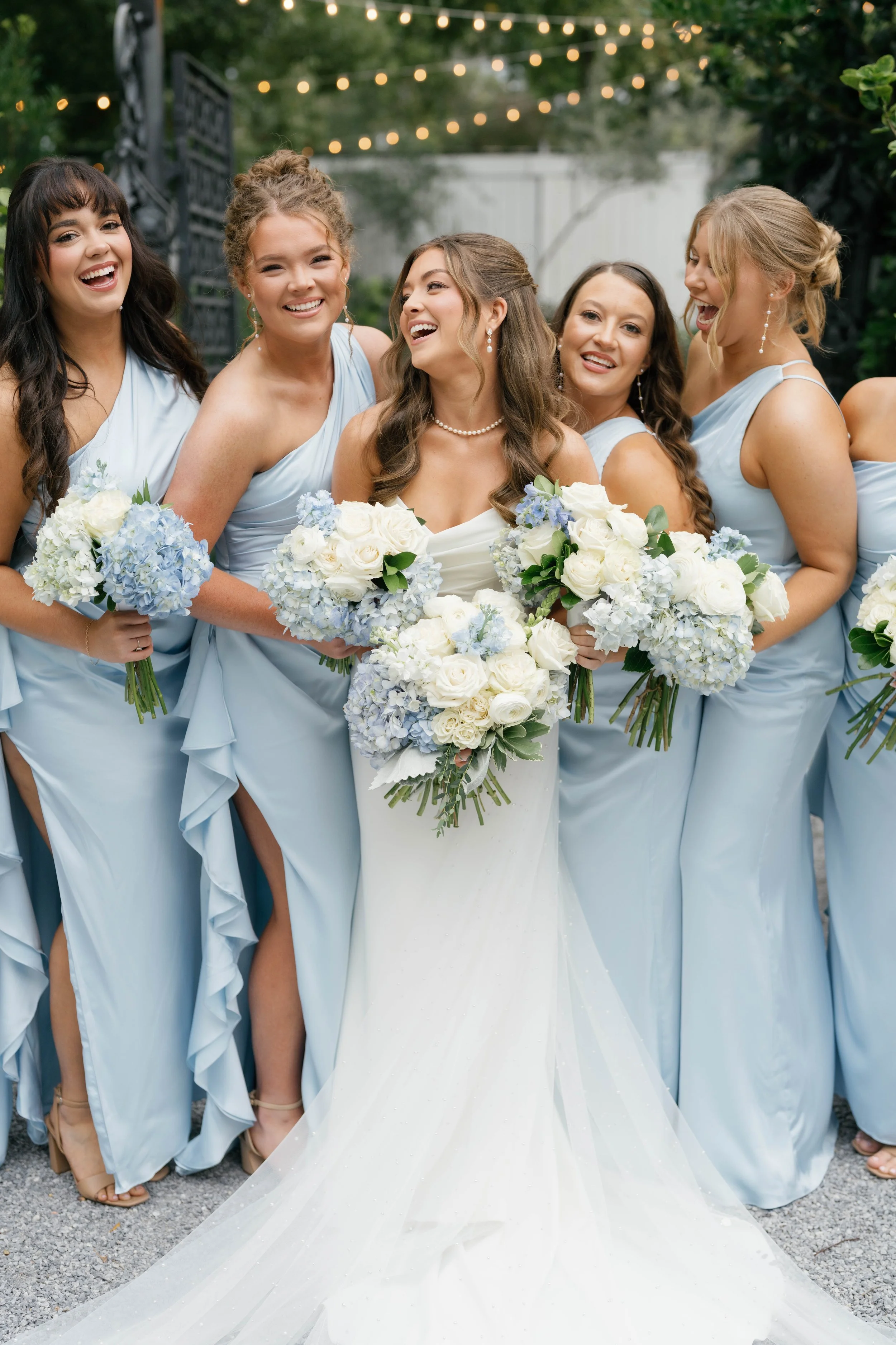 A bride and her bridesmaids standing together outdoors, smiling, with string lights overhead. The bride is wearing a white wedding gown and holding a large bouquet, while the bridesmaids are in matching light blue dresses, each holding smaller bouque