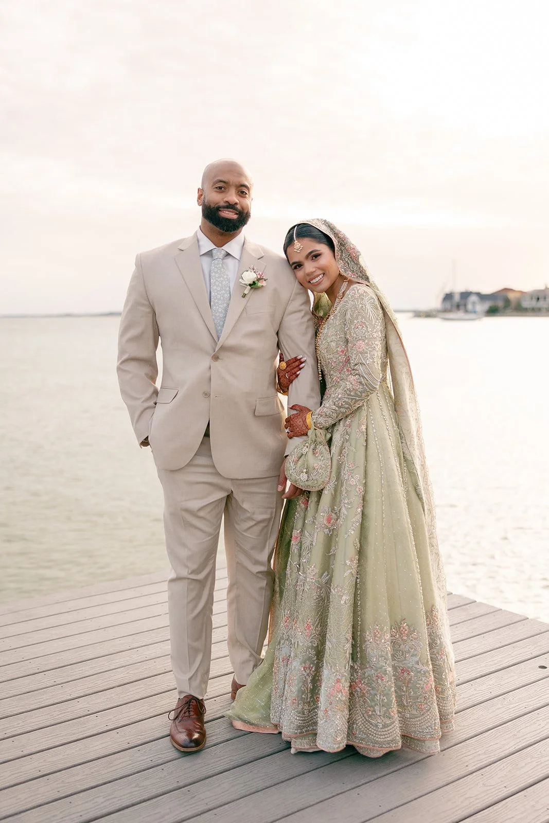 A bride and groom standing on a wooden dock by the water during sunset. The groom is in a light beige suit with a boutonniere, and the bride is wearing an ornate, embroidered traditional dress with jewelry and henna on her hands.