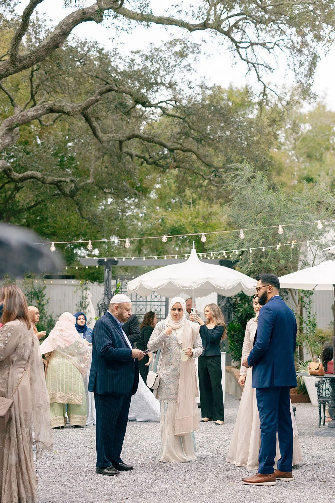 Group of diverse people at an outdoor gathering, with some wearing traditional attire, under string lights and a large white umbrella.