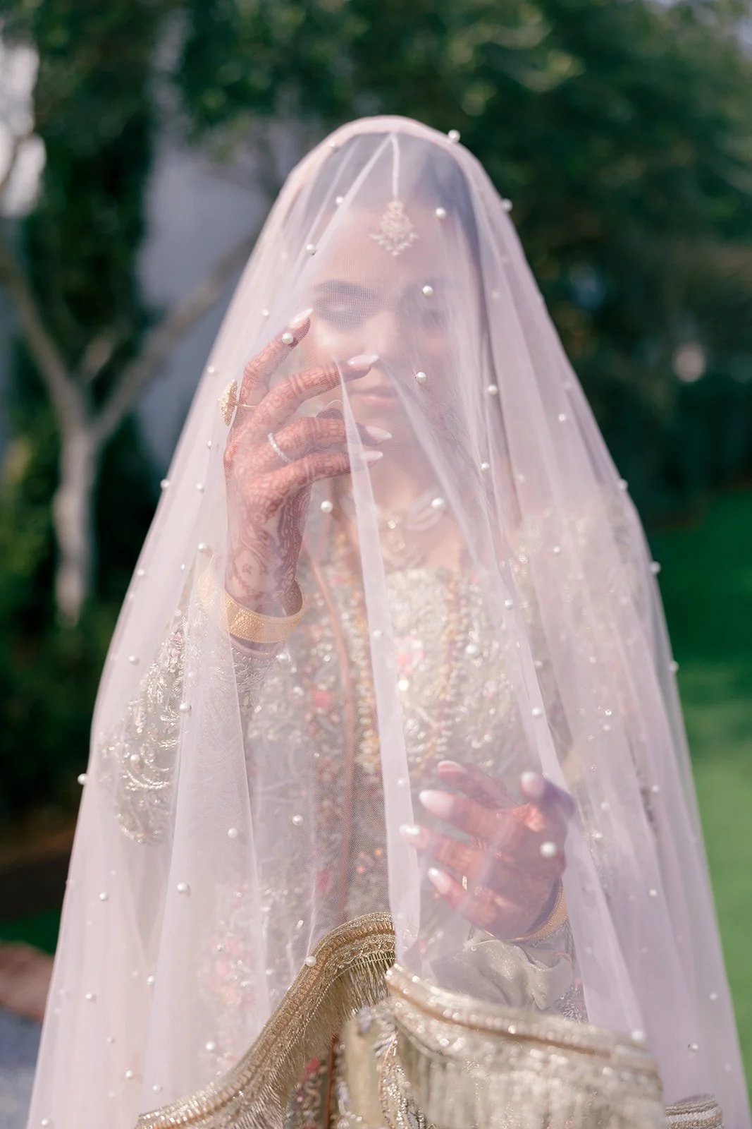A woman dressed in traditional bridal attire, partially obscured by a sheer veil decorated with pearls, outdoors with greenery in the background.