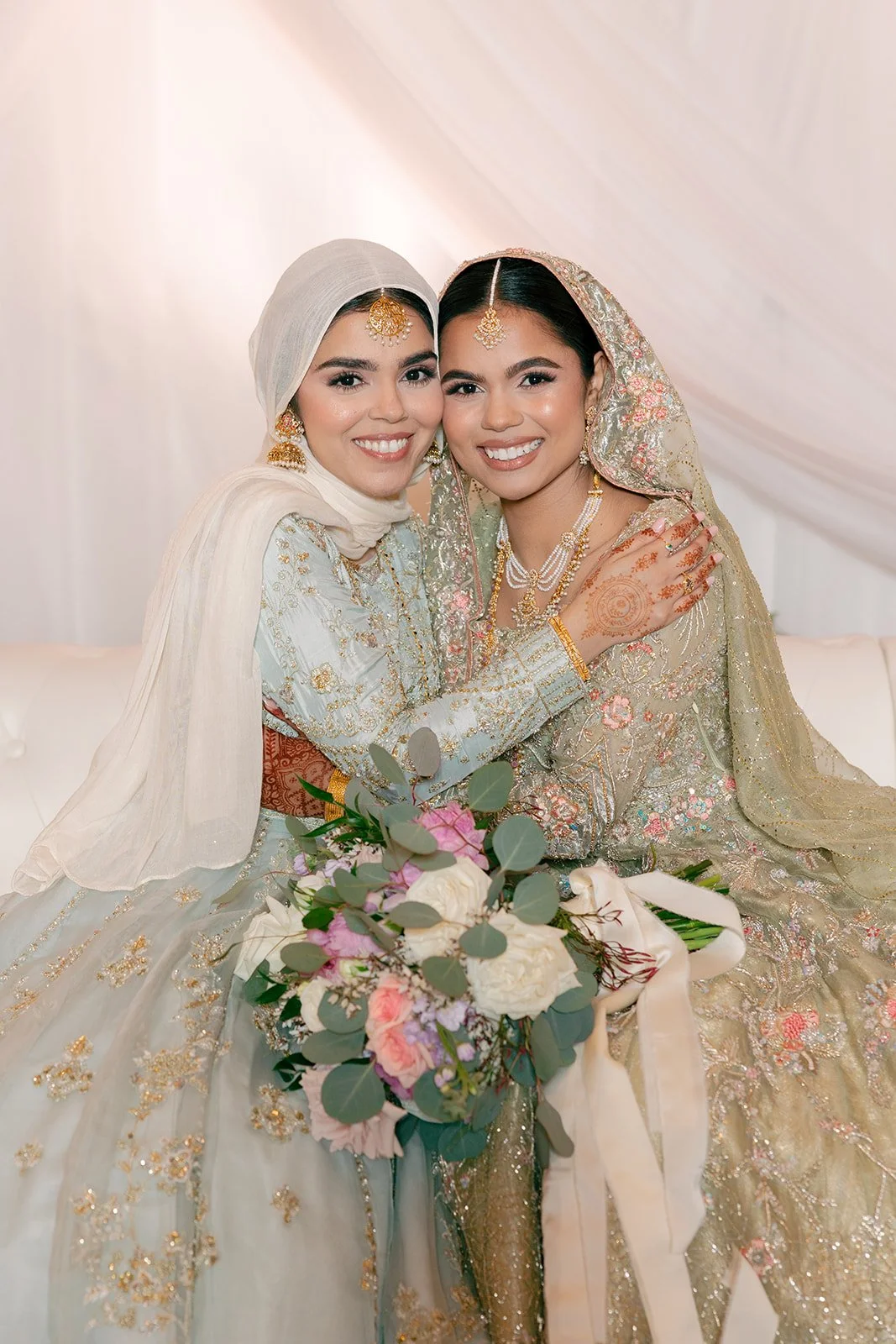 Two women in traditional dresses smiling and hugging, holding a bouquet of flowers, at a wedding celebration.