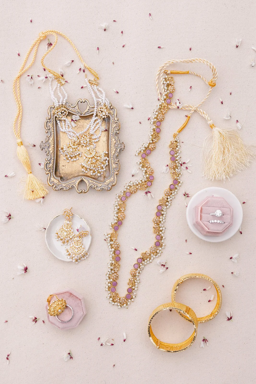 Jewelry collection including necklaces, earrings, bracelets, and rings displayed on a light-colored surface with scattered flower petals.