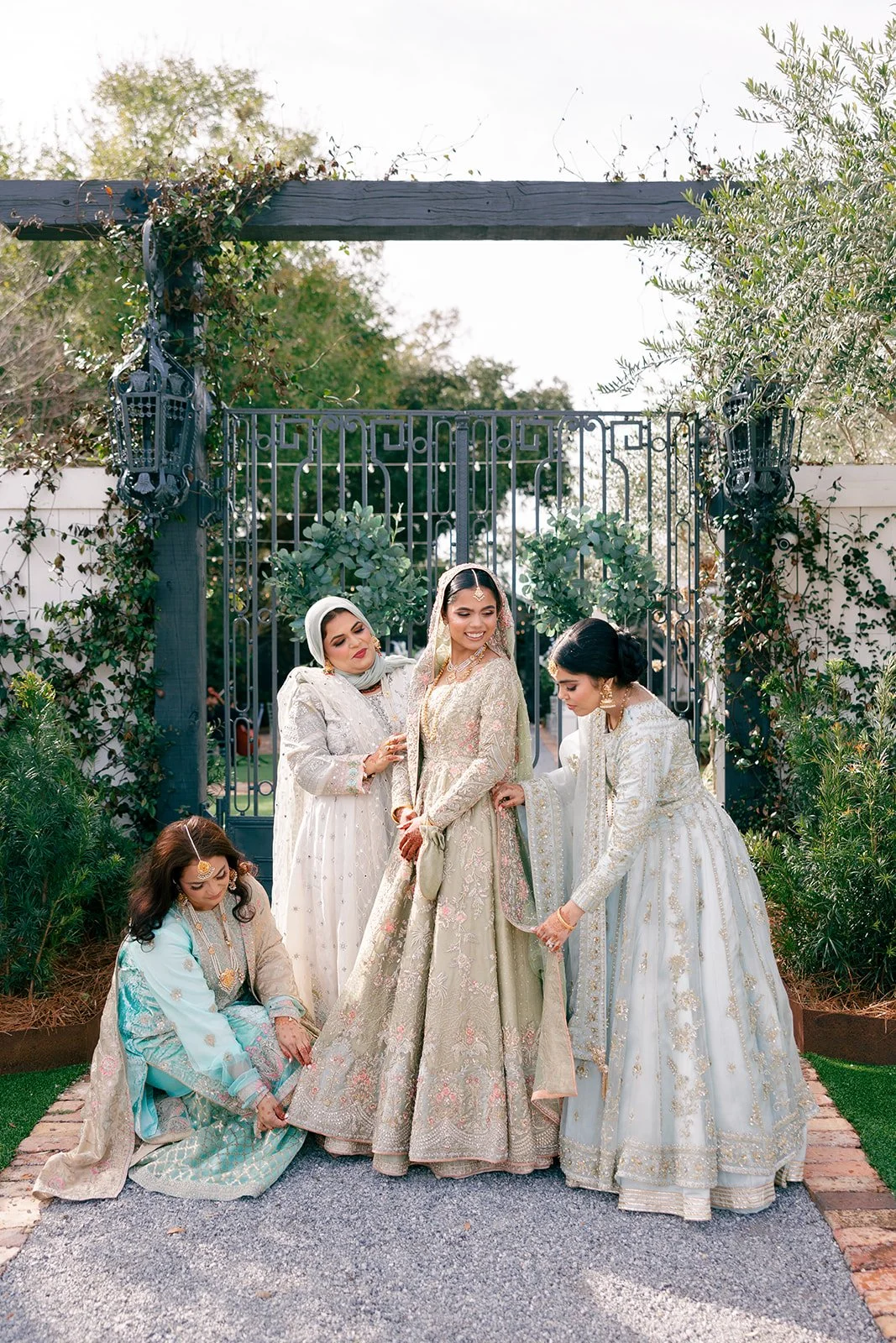Group of women dressed in ornate traditional South Asian attire, gathered outdoors in front of an iron gate, with greenery and trees in the background.