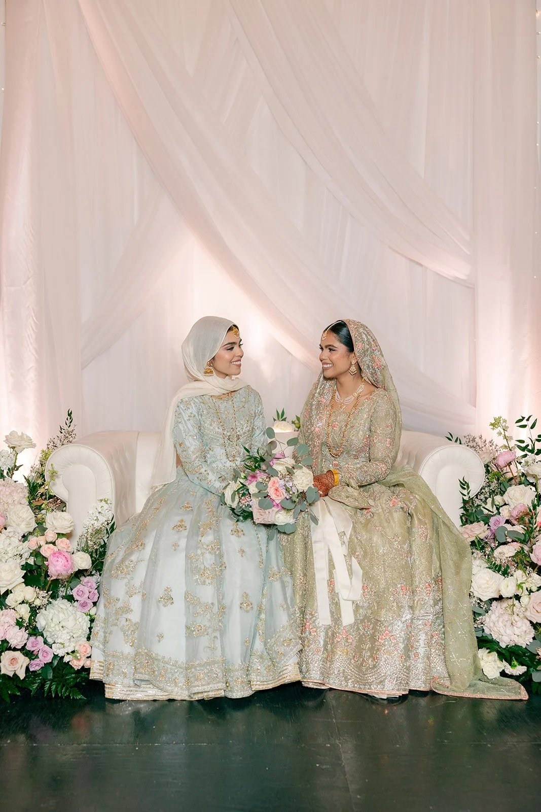 Two women in traditional South Asian wedding attire sitting on a white sofa, smiling at each other, with floral arrangements surrounding them and a pink draped backdrop behind.