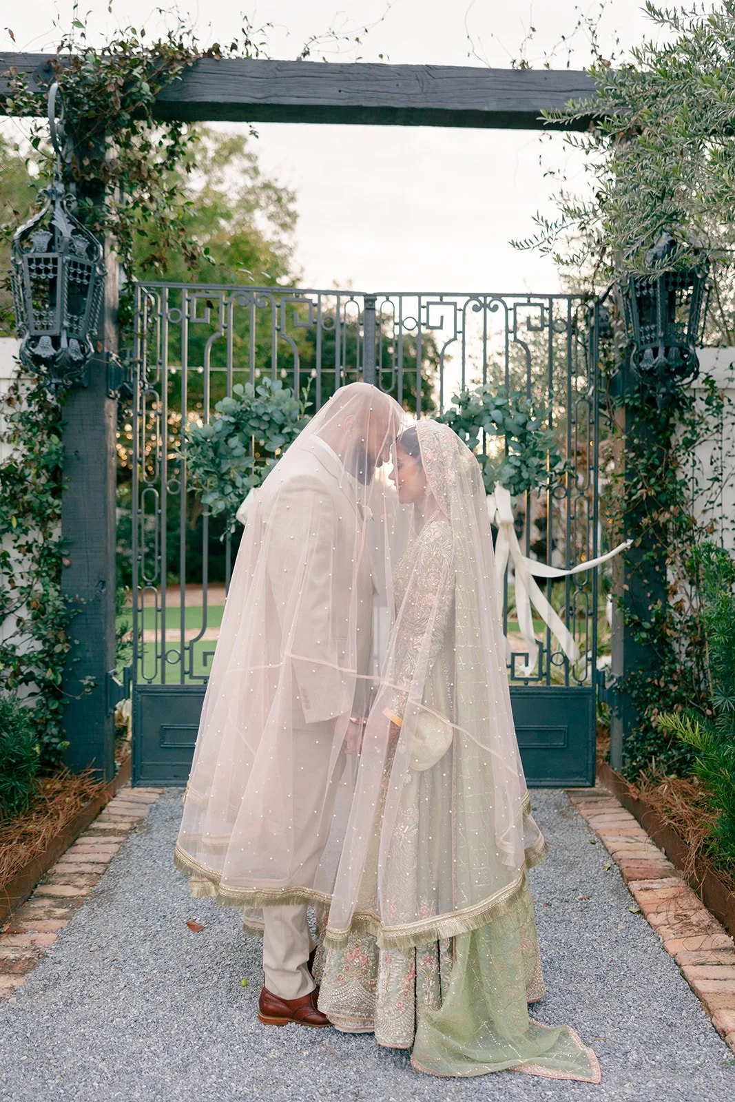 A bride and groom dressed in traditional Indian wedding attire standing close behind a wedding veil, touching foreheads, outside in front of a decorative iron gate surrounded by greenery.