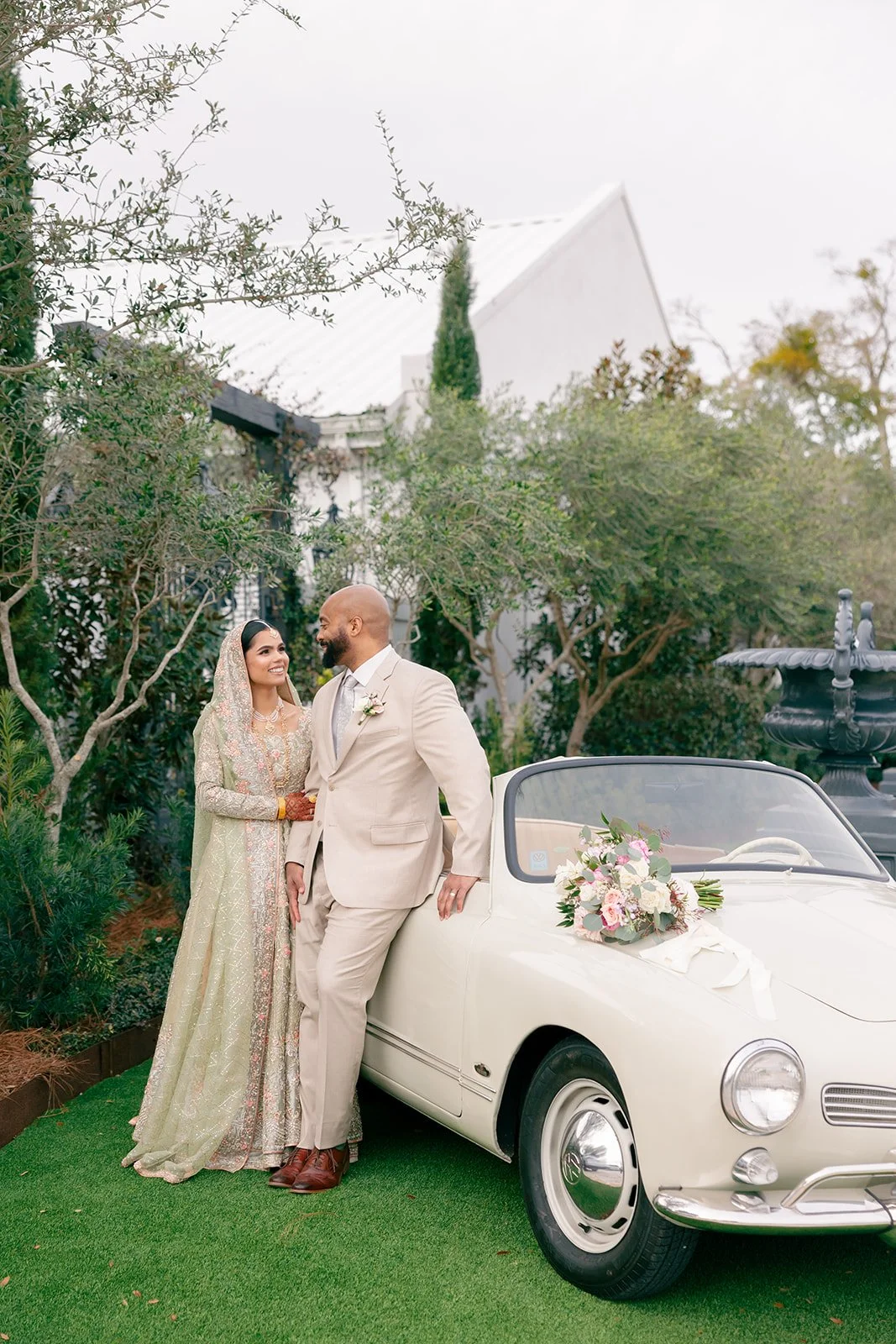 A bride and groom standing next to a vintage white convertible car with a floral arrangement on the hood. The bride wears a traditional dress with a veil, and the groom wears a tan suit. They are outdoors, surrounded by greenery and trees, and appear
