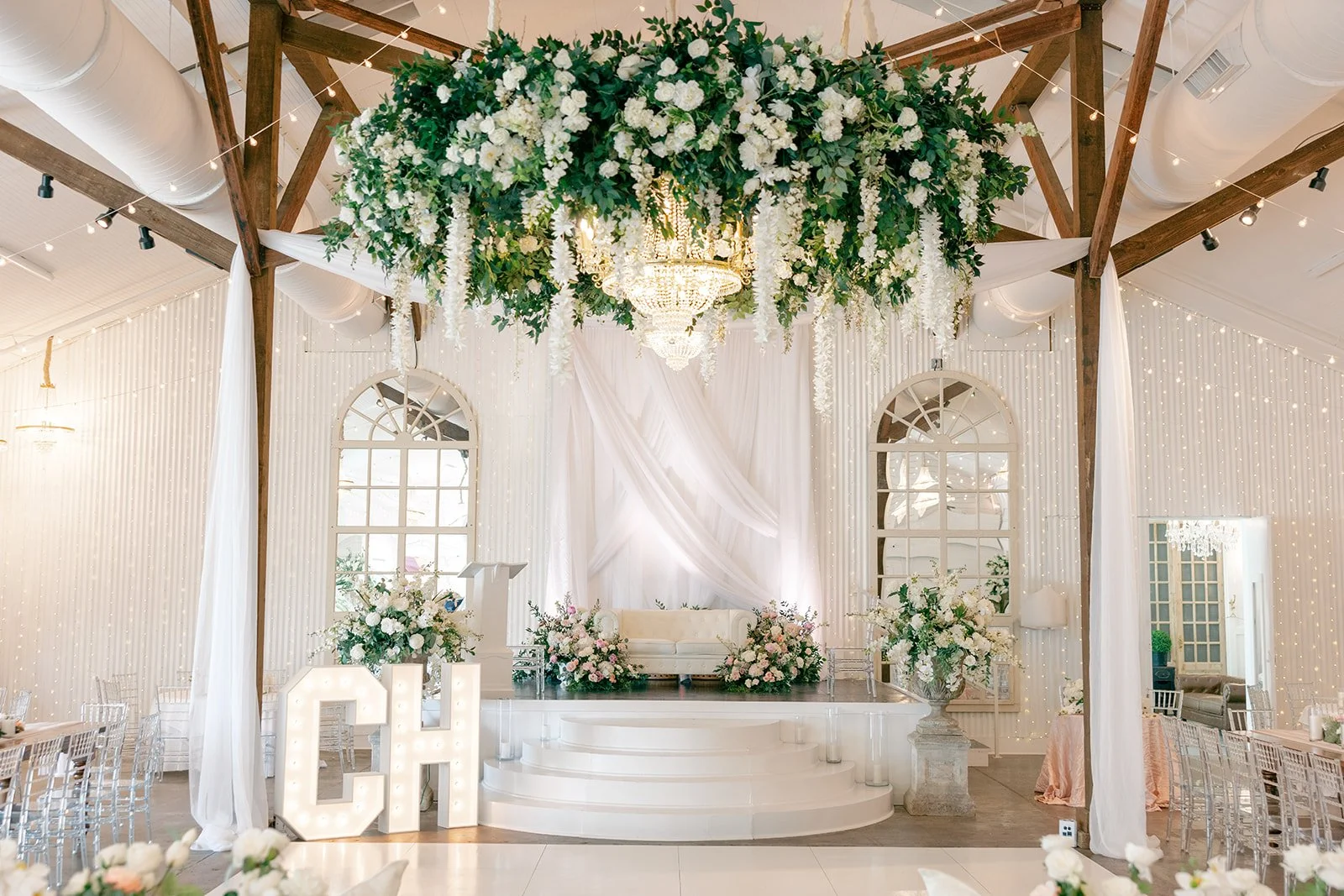 Elegant Pensacola  wedding ceremony setup with a white and gold theme, featuring a stage with floral arrangements, a crystal chandelier, and draped white fabric backdrop.