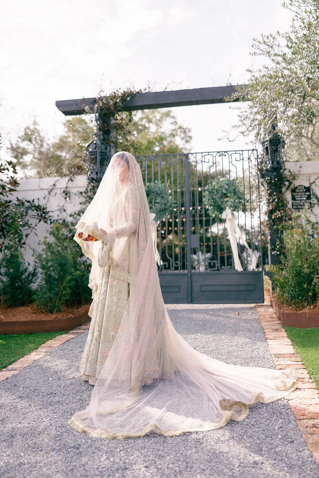 A bride in an ornate beige dress with a sheer veil stands on a gravel pathway near an iron gate covered in greenery, with trees in the background of a Pensacola Wedding Venue.