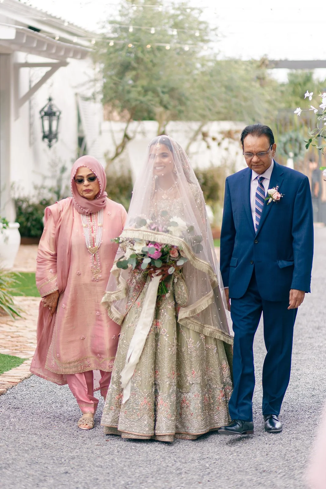 A bride in a traditional wedding gown and veil holding a bouquet, walking outdoors with two older adults, one woman dressed in pink traditional attire and sunglasses, and one man in a blue suit with a tie.