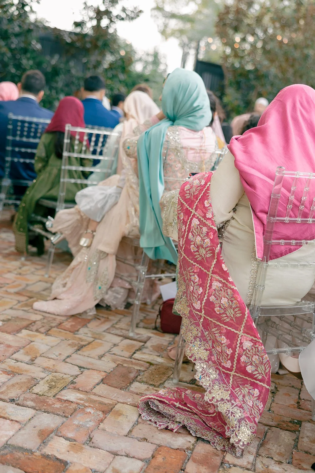 People sitting on clear chairs outside, wearing colorful traditional clothing and headscarves, on a brick patio with trees in the background.
