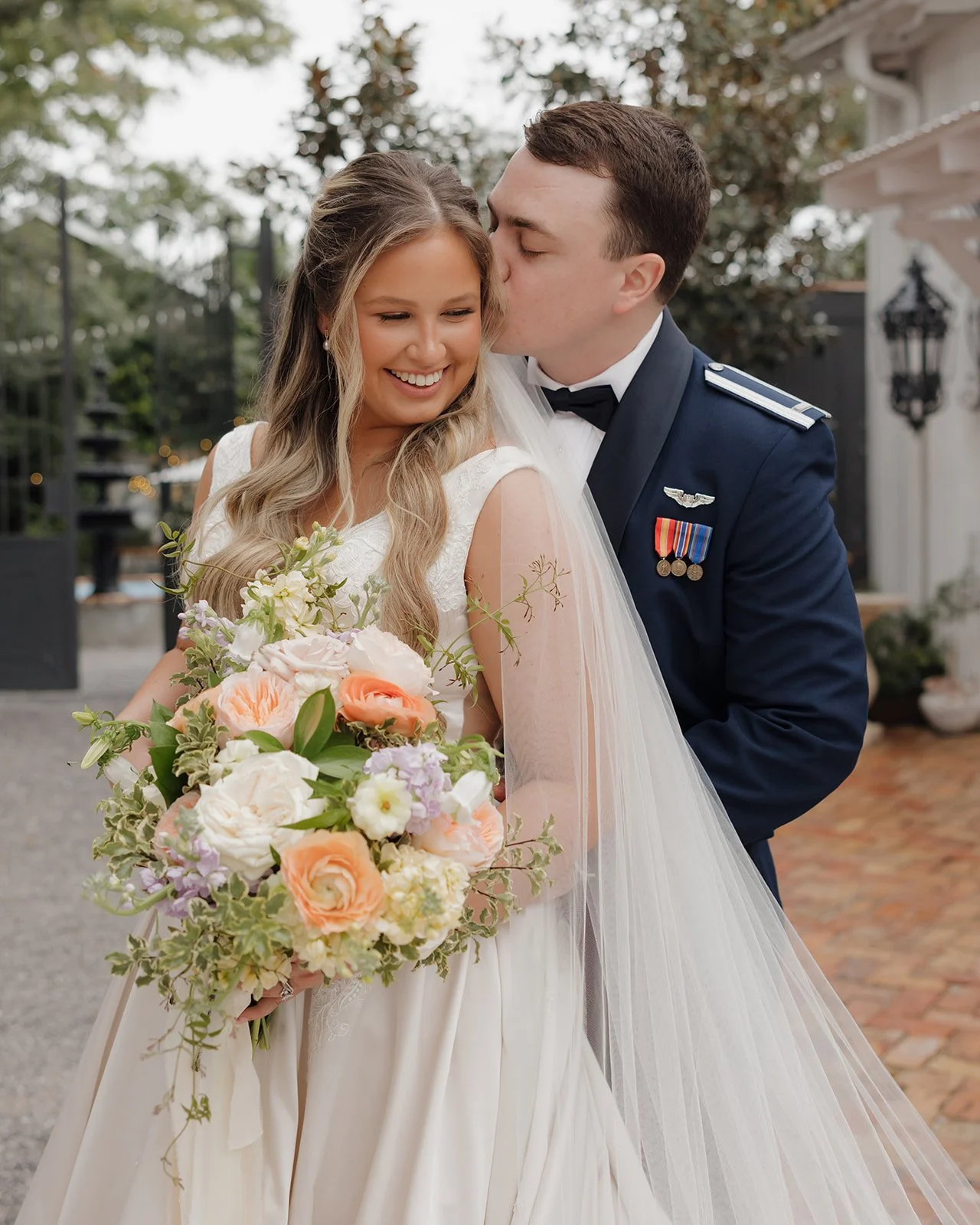 A newlywed Pensacola couple, the bride in a white wedding dress holding a bouquet of flowers, the groom in a military uniform, sharing a kiss outdoors during daytime.