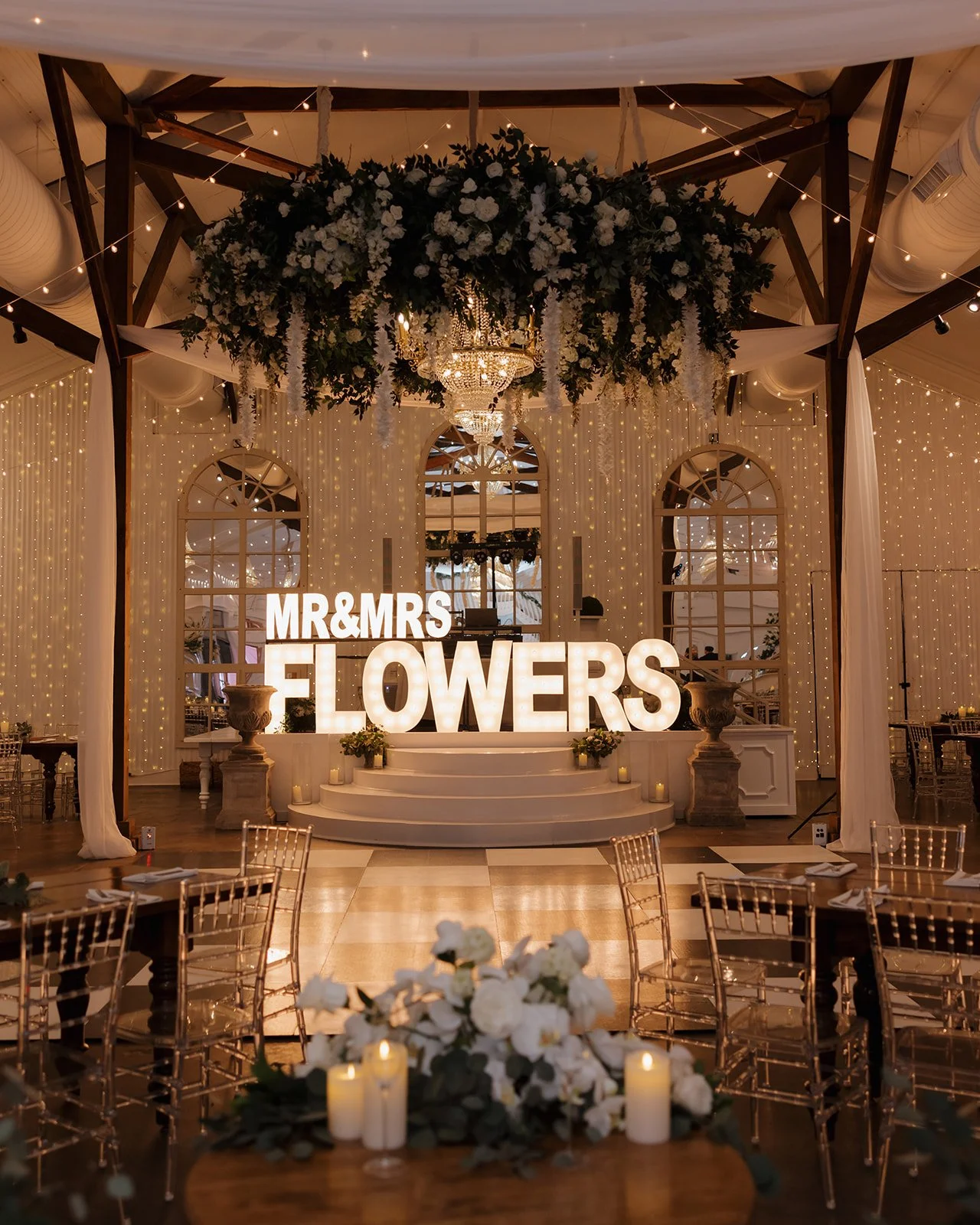 Wedding reception decorated with white flowers, candles, and fairy lights. Large illuminated sign reads 'MR & MRS FLOWERS' on a small stage with steps, flanked by large planter urns. Round tables with chairs are set for guests, with floral centerpiec
