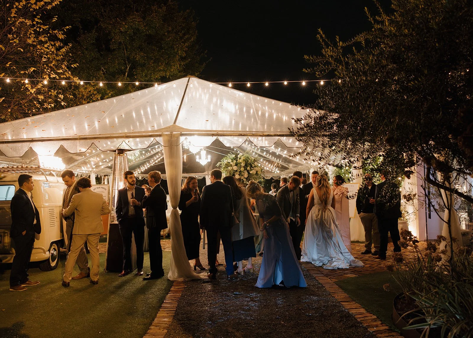 People gathering under a lit-up white tent at night, with string lights overhead, at an outdoor event or wedding reception.