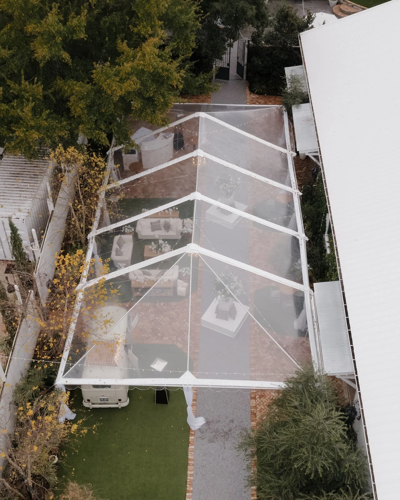An outdoor patio covered by a transparent tent with white framing. The patio contains outdoor furniture, including sofas and a table, and is surrounded by trees and nearby buildings.
