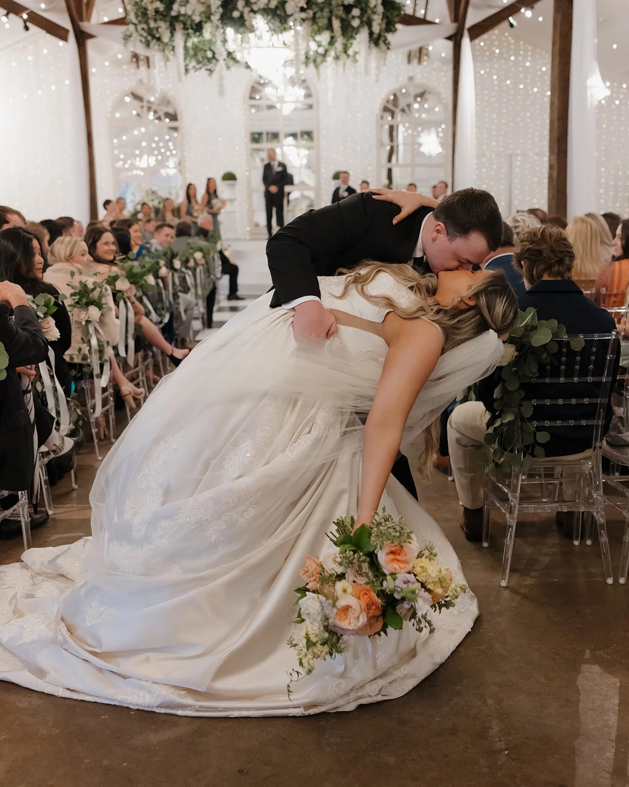 A Pensacola Bride and groom share a kiss during their wedding ceremony, with the groom leaning over the bride who is in a white wedding dress holding a bouquet of flowers. Guests are seated around them in a decorated venue.