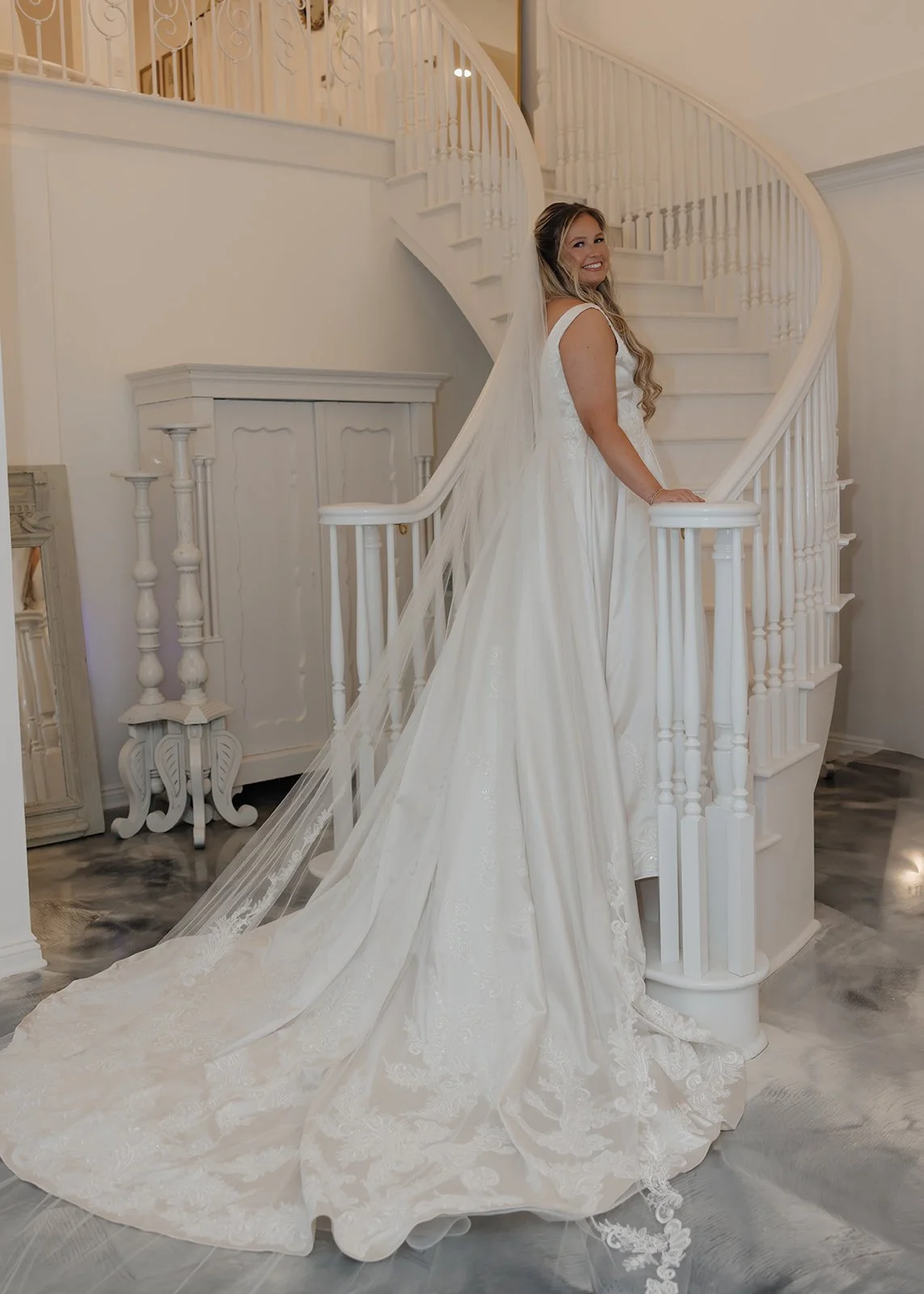 A bride in a white wedding dress with a long train and veil, standing on a staircase in a white interior of a Pensacola Wedding Venue.