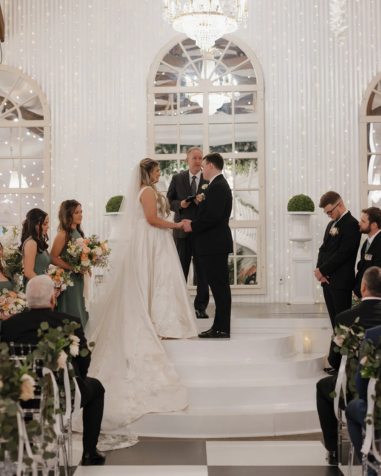 A Pensacola wedding ceremony with a bride and groom holding hands on stage, surrounded by bridesmaids and groomsmen, with an officiant and guests in a decorated indoor venue with fairy lights and chandeliers.