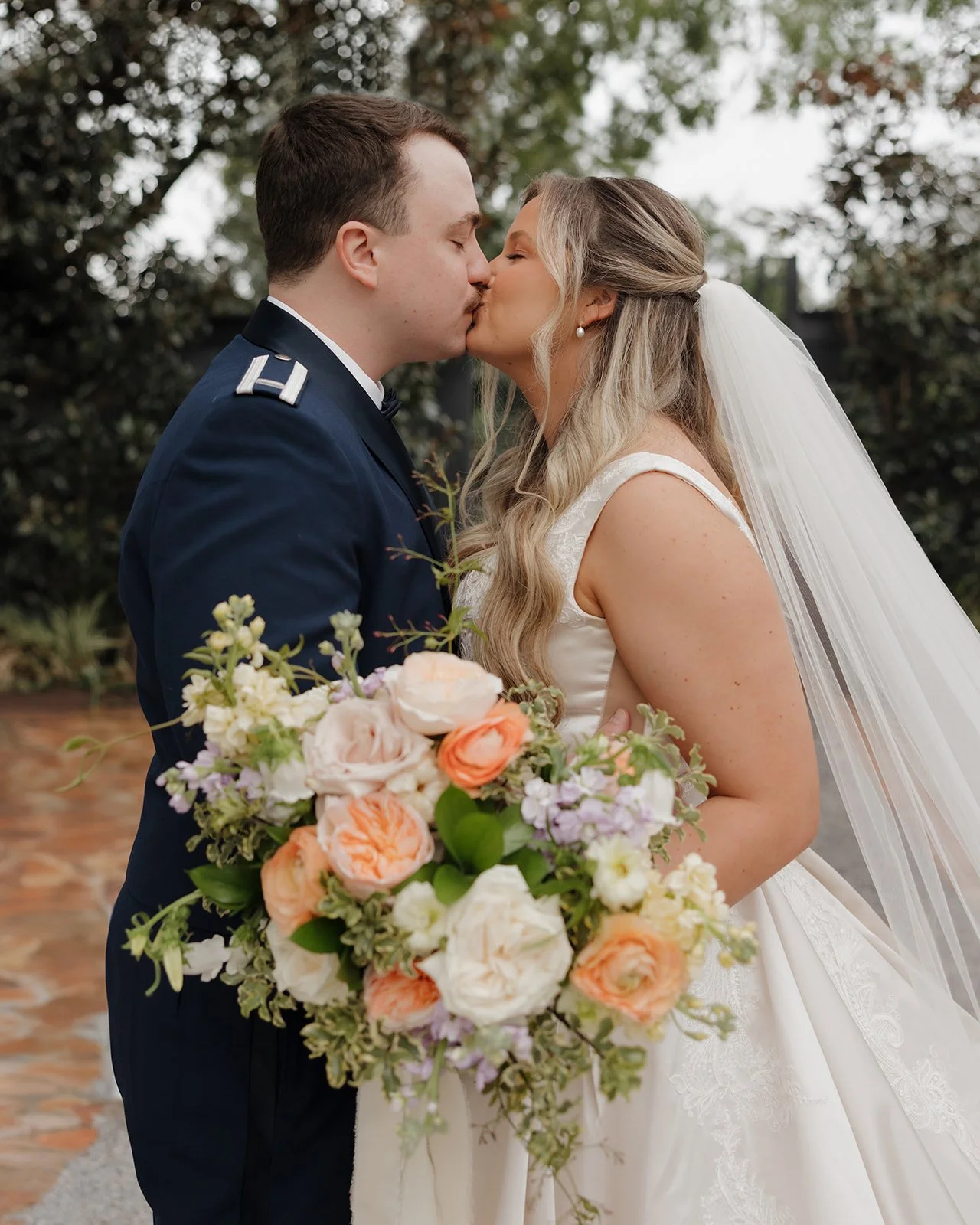 A Pensacola bride and groom kissing outdoors while holding a large bouquet of white, peach, and lavender flowers.