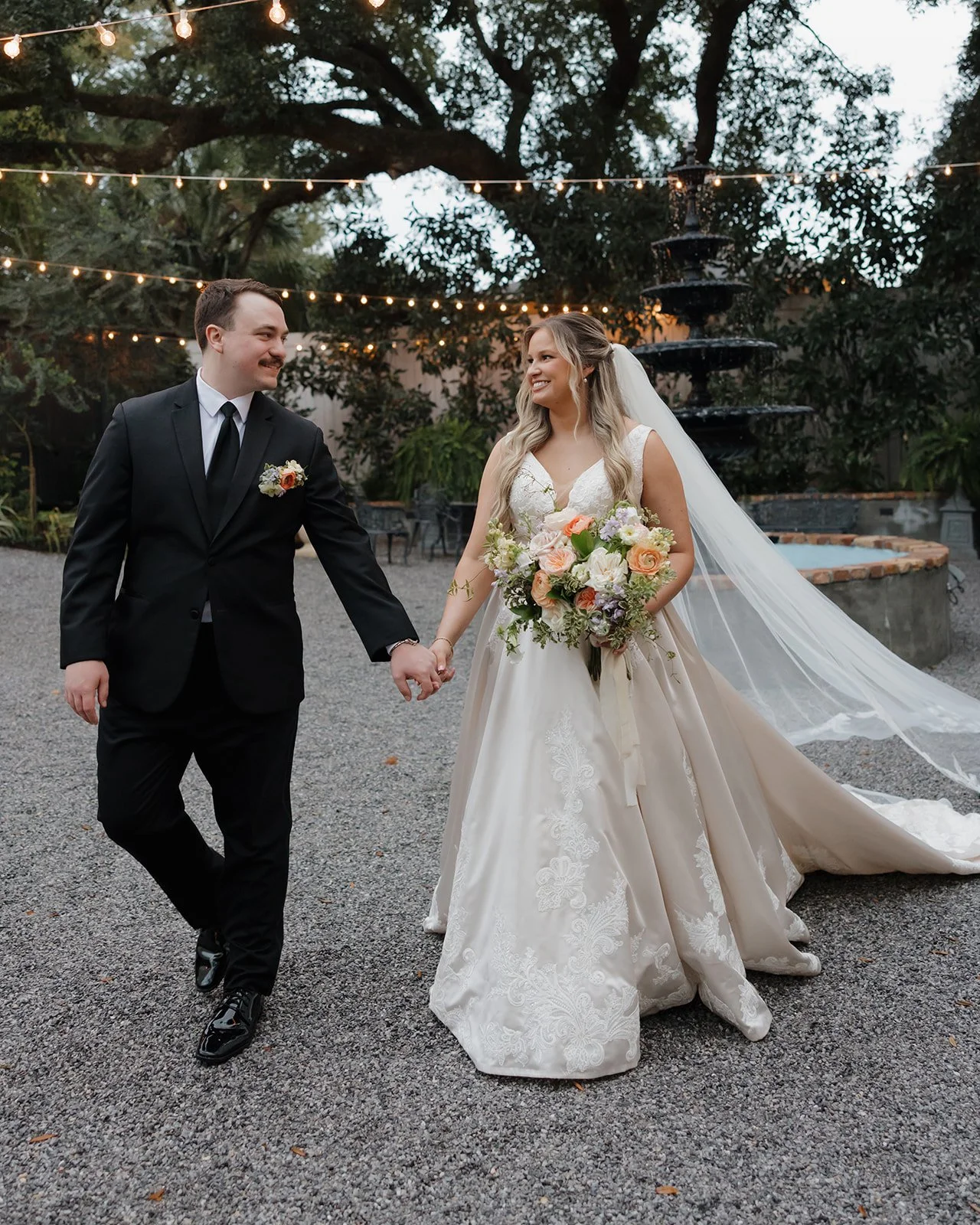 A Pensacola bride and groom holding hands and smiling at each other outdoors at sunset, with string lights overhead, a fountain in the background, and trees surrounding them.